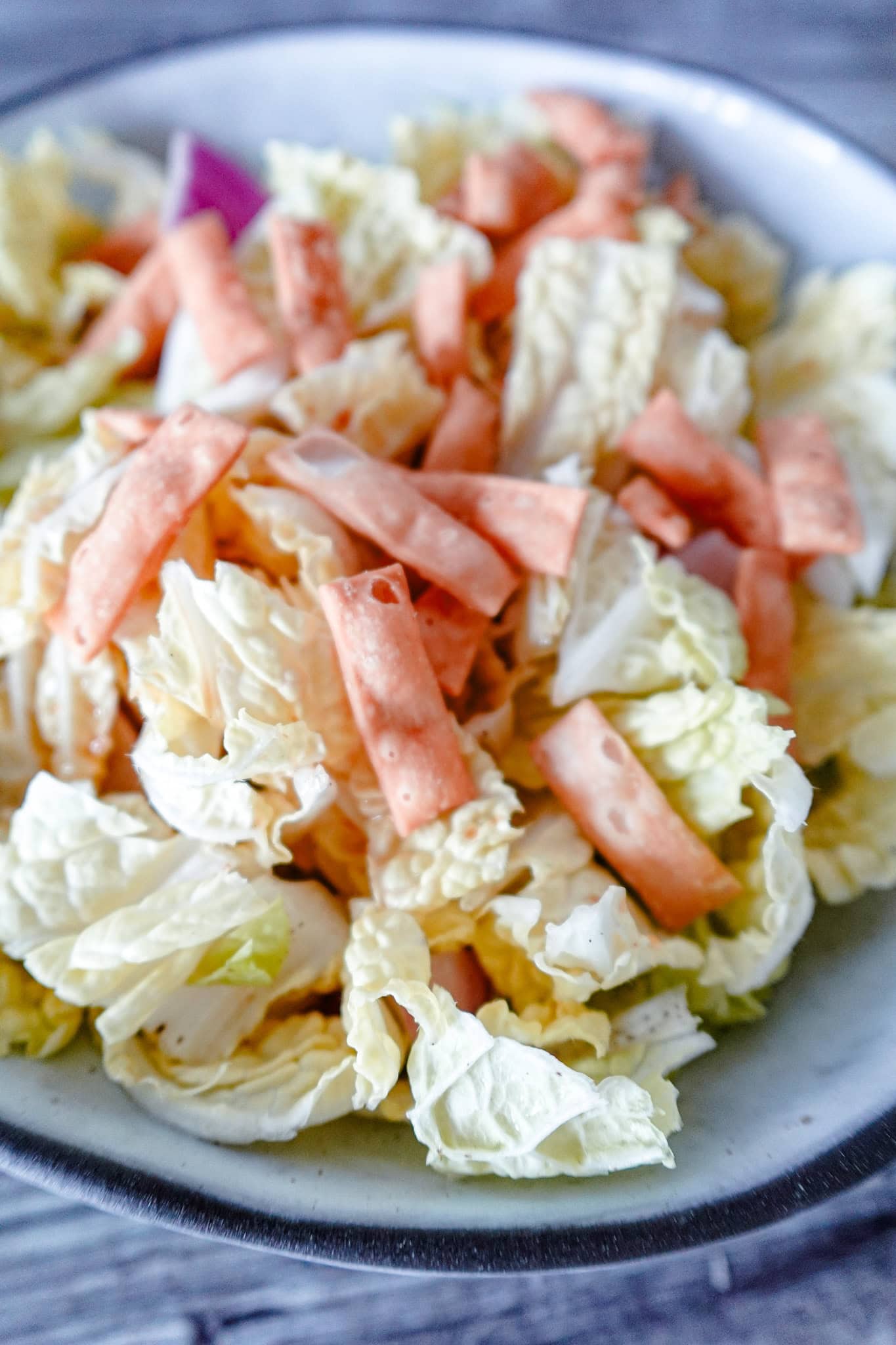 A vertical shot of napa cabbage salad in a white bowl, highlighting crisp cabbage leaves, carrot strips, red onion, and crunchy wonton strips.