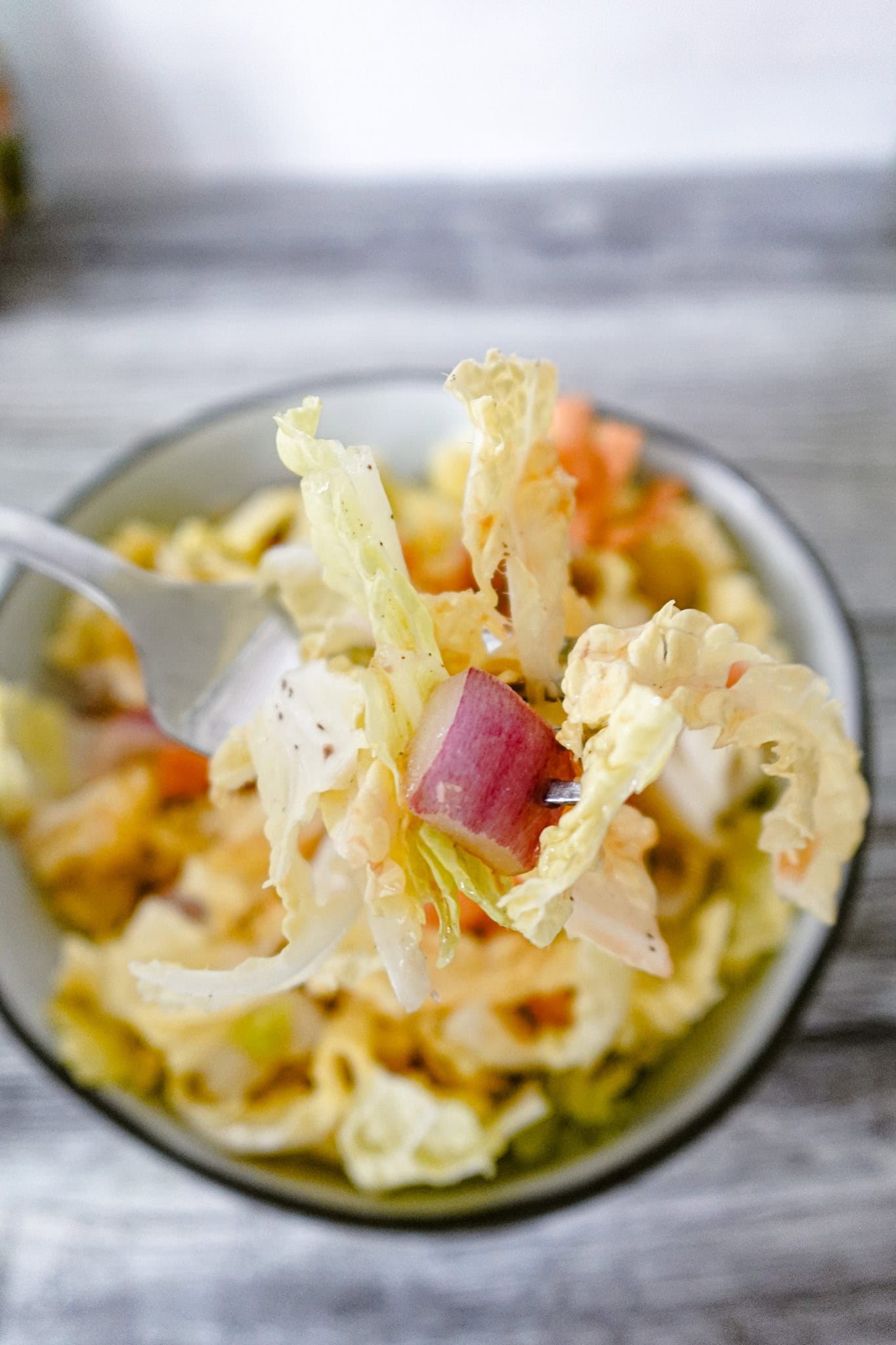A fork holding a bite of napa cabbage salad, featuring a piece of cabbage and a chunk of red onion, with the bowl of salad blurred in the background.