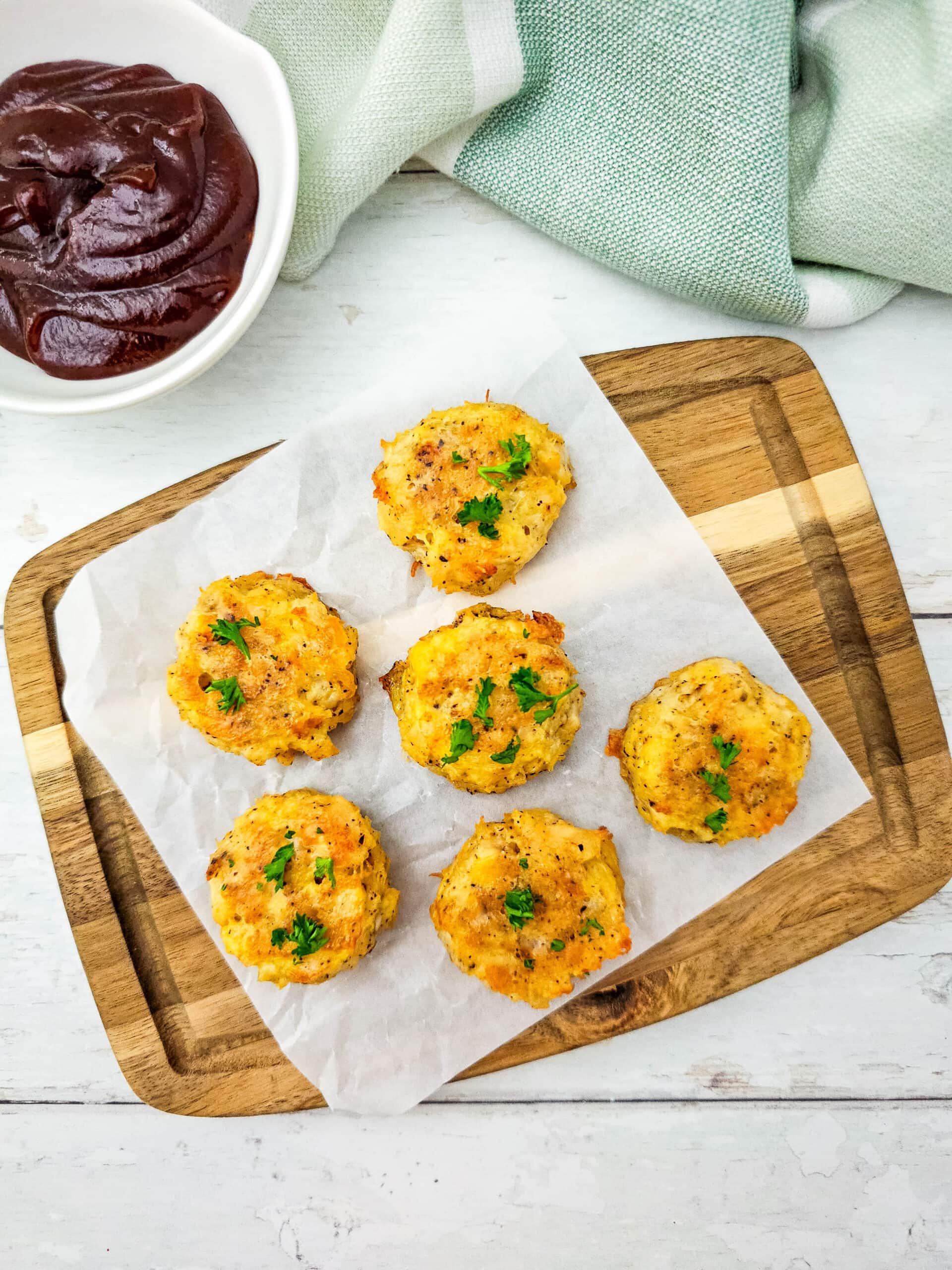 Six baked low carb chicken nuggets garnished with parsley, served on parchment paper on a wooden board with barbecue dipping sauce in the background.