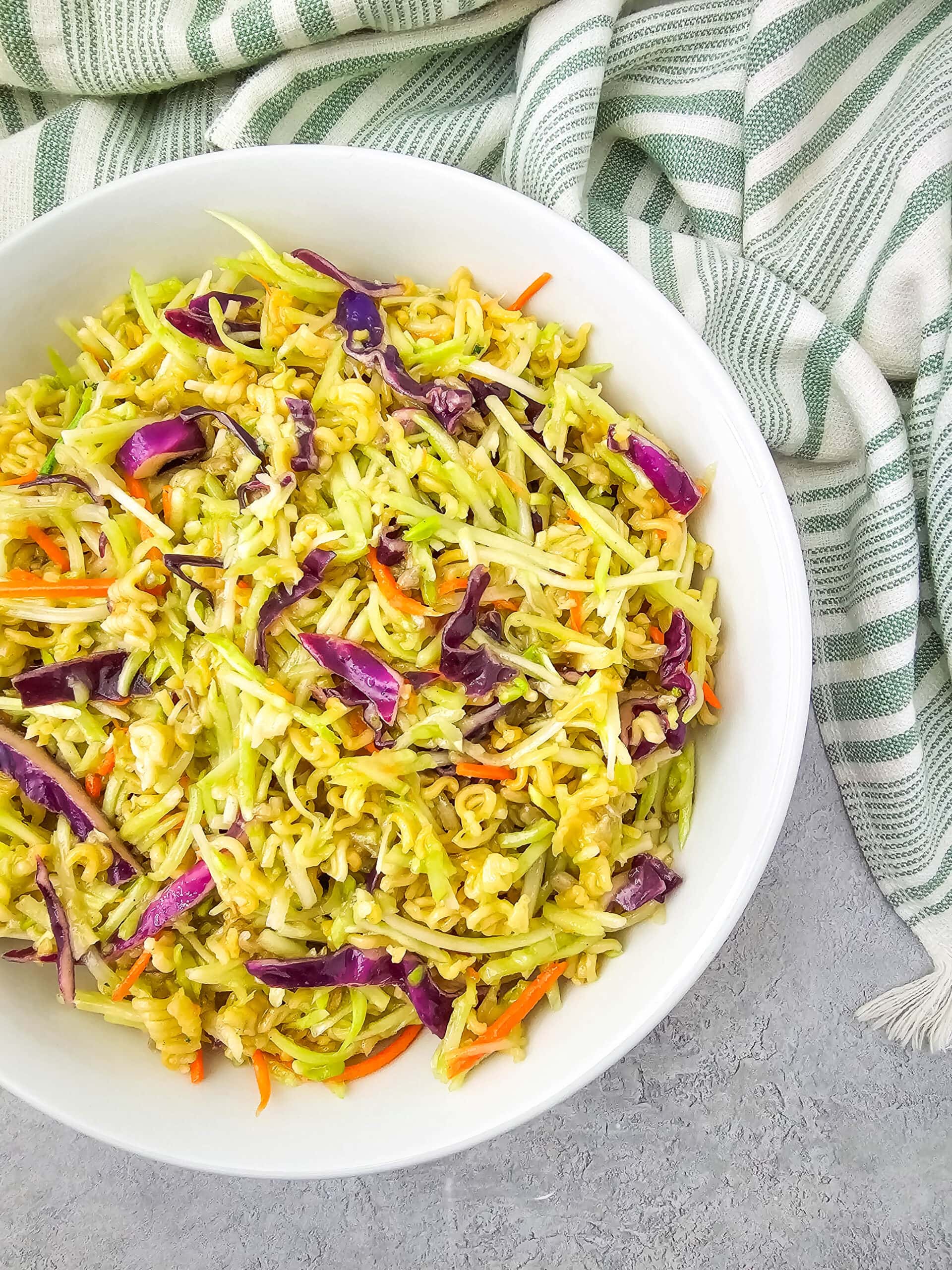 Overhead view of Ramen Noodle Broccoli Slaw Salad in a large white serving bowl, surrounded by a soft green and white striped kitchen towel.