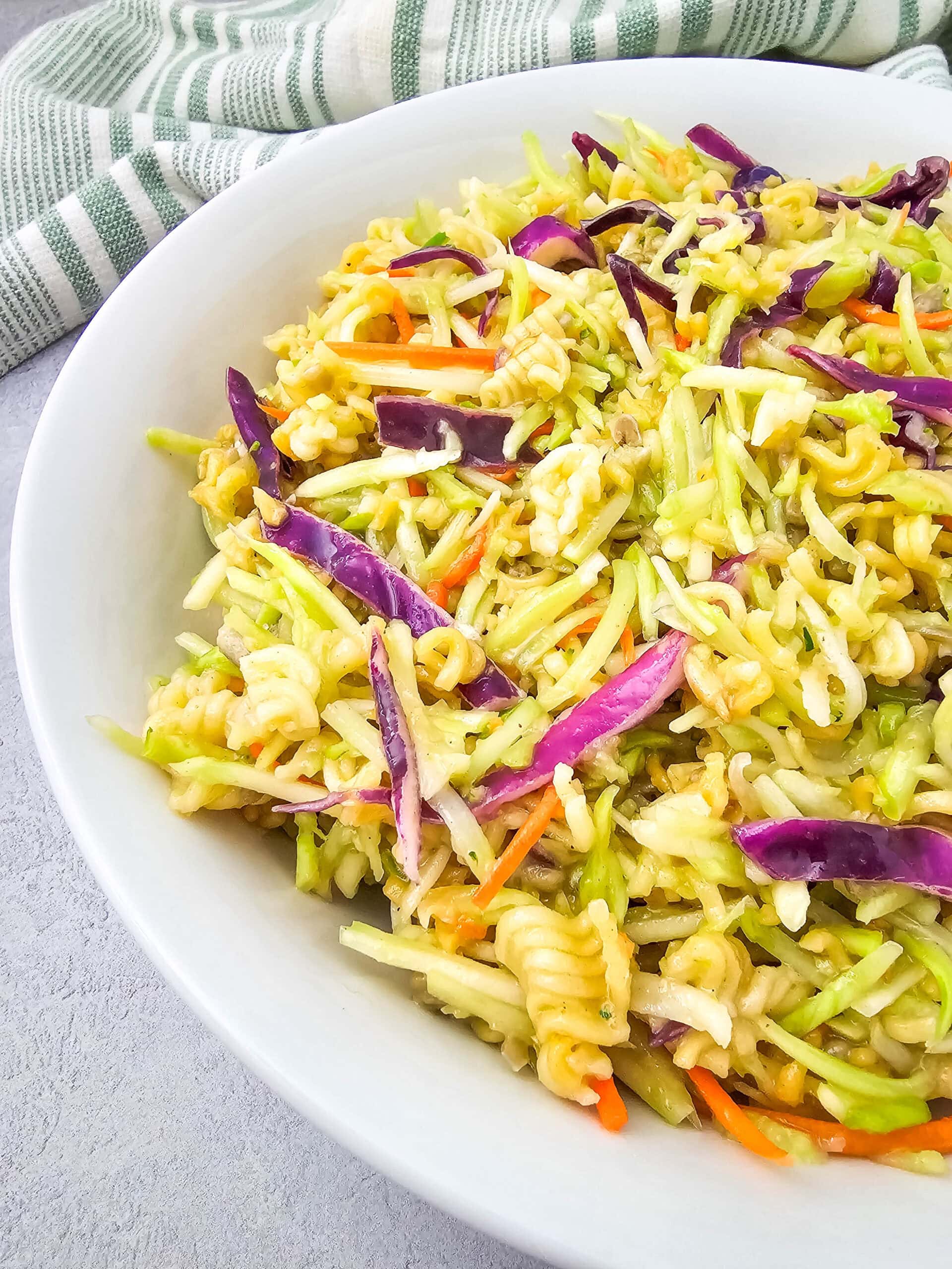 A close-up view of Ramen Noodle Broccoli Slaw Salad in a white bowl, showing colorful shredded cabbage, carrots, and broken ramen noodles.