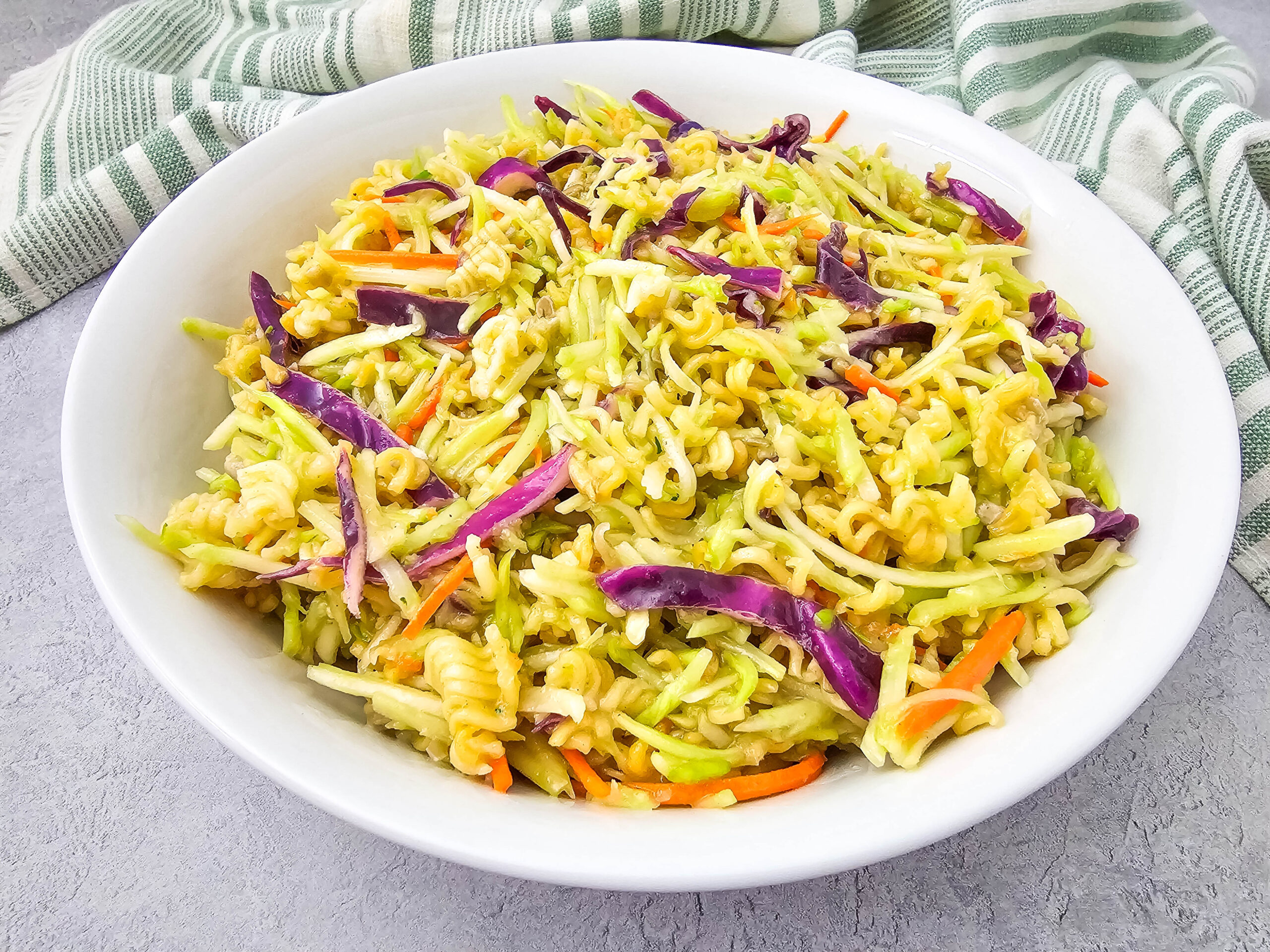 Overhead view of a large white bowl filled with broccoli slaw salad mixed with crunchy ramen noodles, red cabbage, and carrots, next to a green-striped towel.