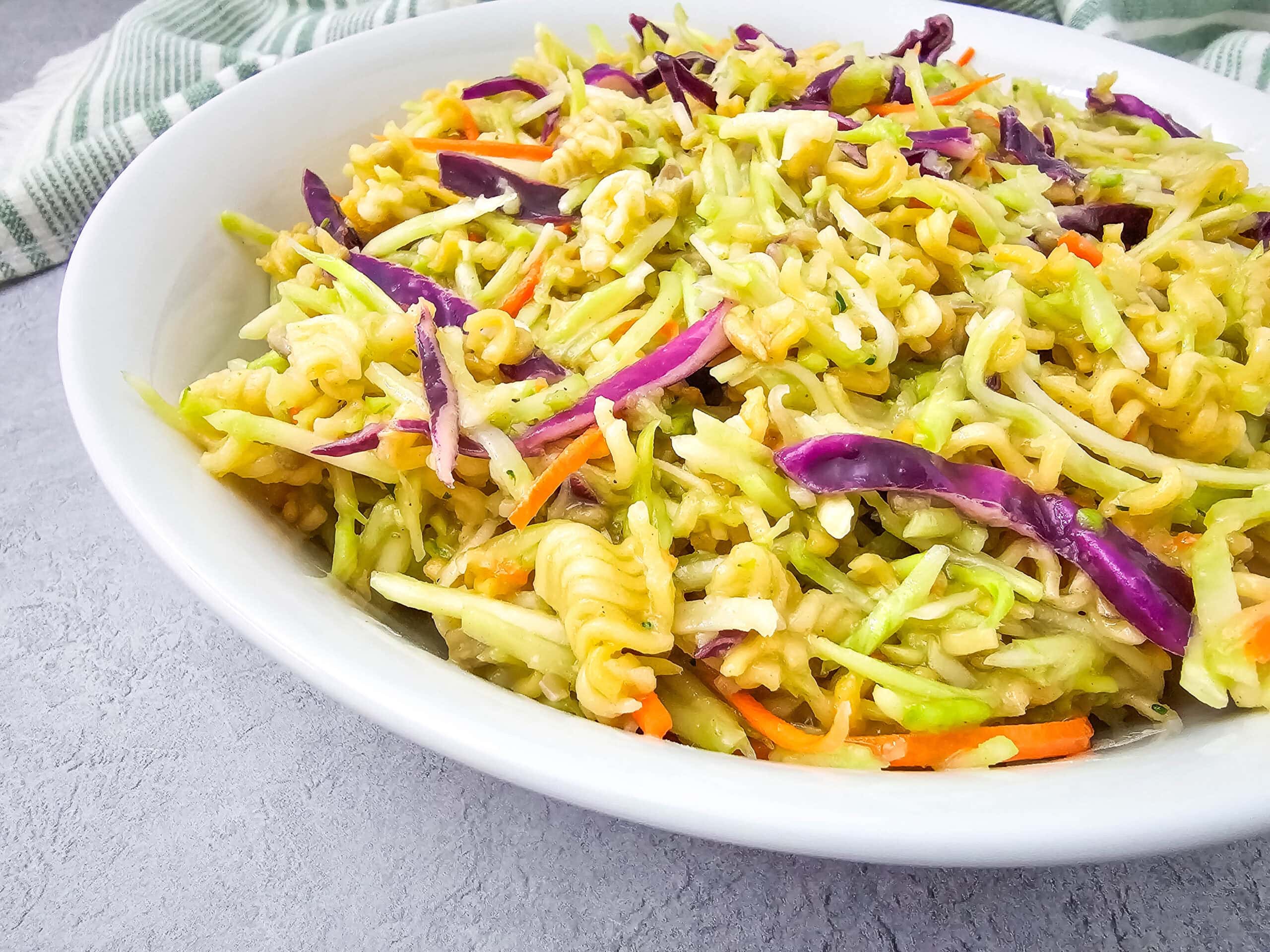 A side view of broccoli slaw salad tossed with ramen noodles, red cabbage, and carrots in a white bowl on a light grey surface.