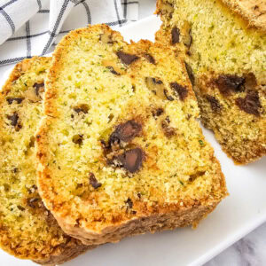 Overhead view of a baked zucchini bread loaf and two slices on a white serving plate, set on a marble surface with a checkered kitchen towel in the background.