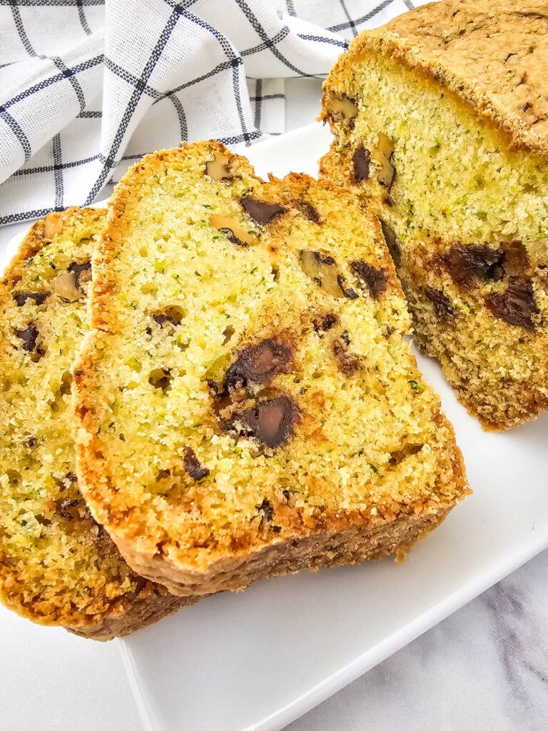 Overhead view of a baked zucchini bread loaf and two slices on a white serving plate, set on a marble surface with a checkered kitchen towel in the background.