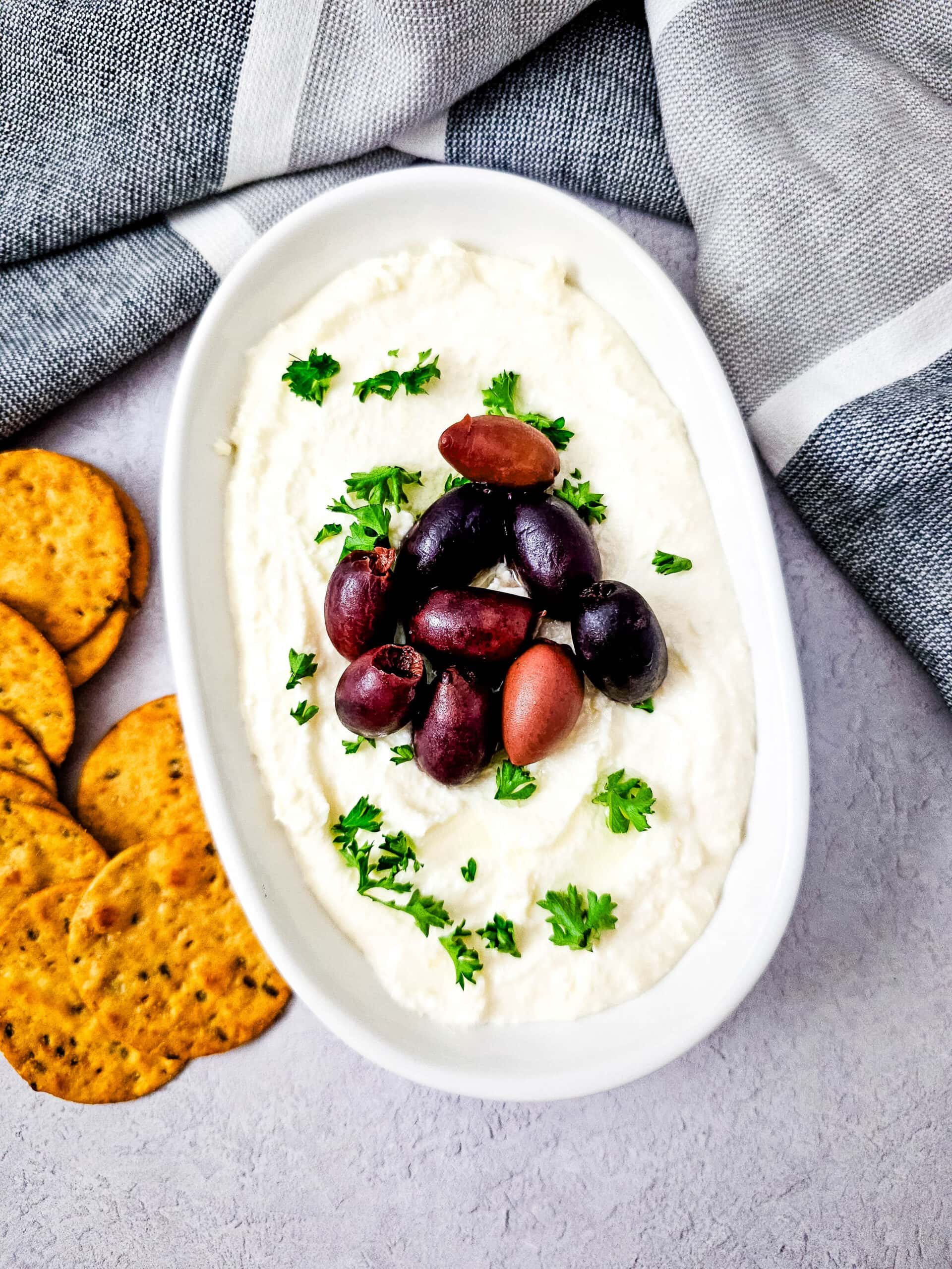 Overhead view of whipped feta dip in a white dish, decorated with olives and parsley, with crackers to the side on a light surface.