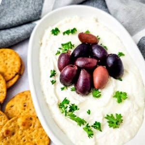 Oval dish of creamy whipped feta dip topped with Kalamata olives and fresh parsley, served with crackers on the side.