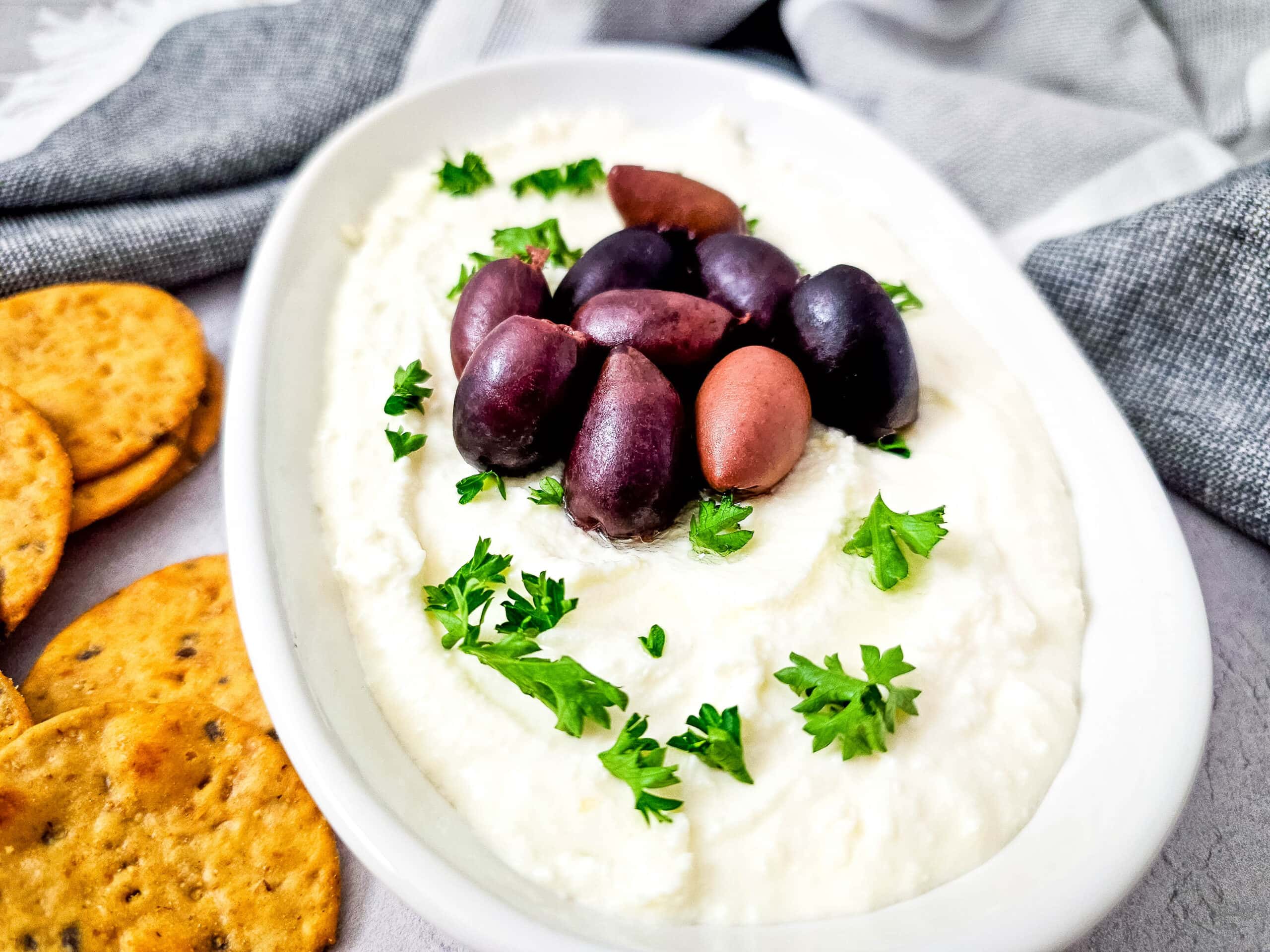 Angled view of whipped feta dip topped with black and red olives, surrounded by sprigs of parsley and served with crackers.