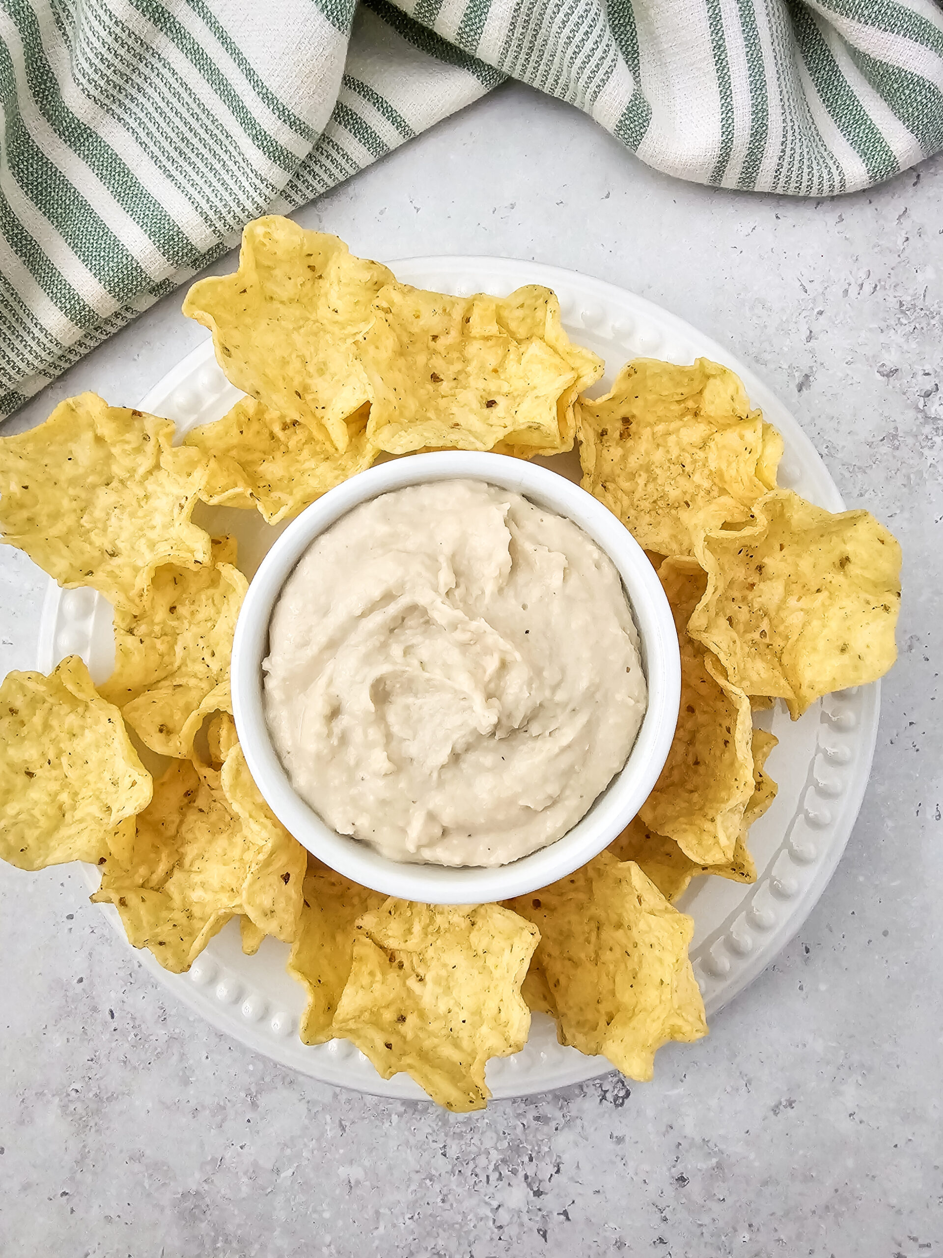 Bowl of creamy white bean dip surrounded by yellow tortilla scoop chips on a white plate, with a green-striped towel in the background.