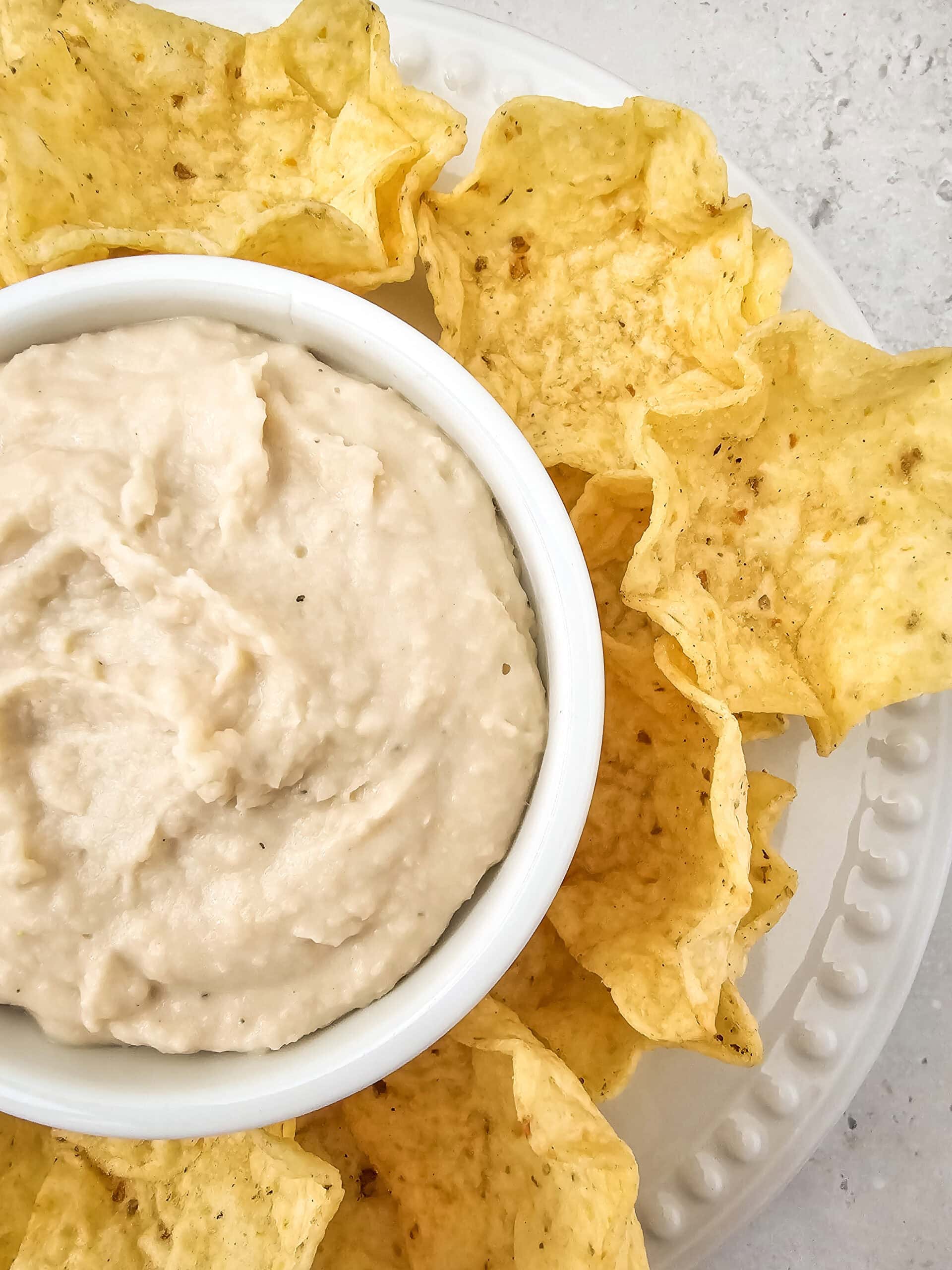 Close-up of smooth, velvety white bean dip in a small white ramekin, encircled by tortilla chips on a serving plate.