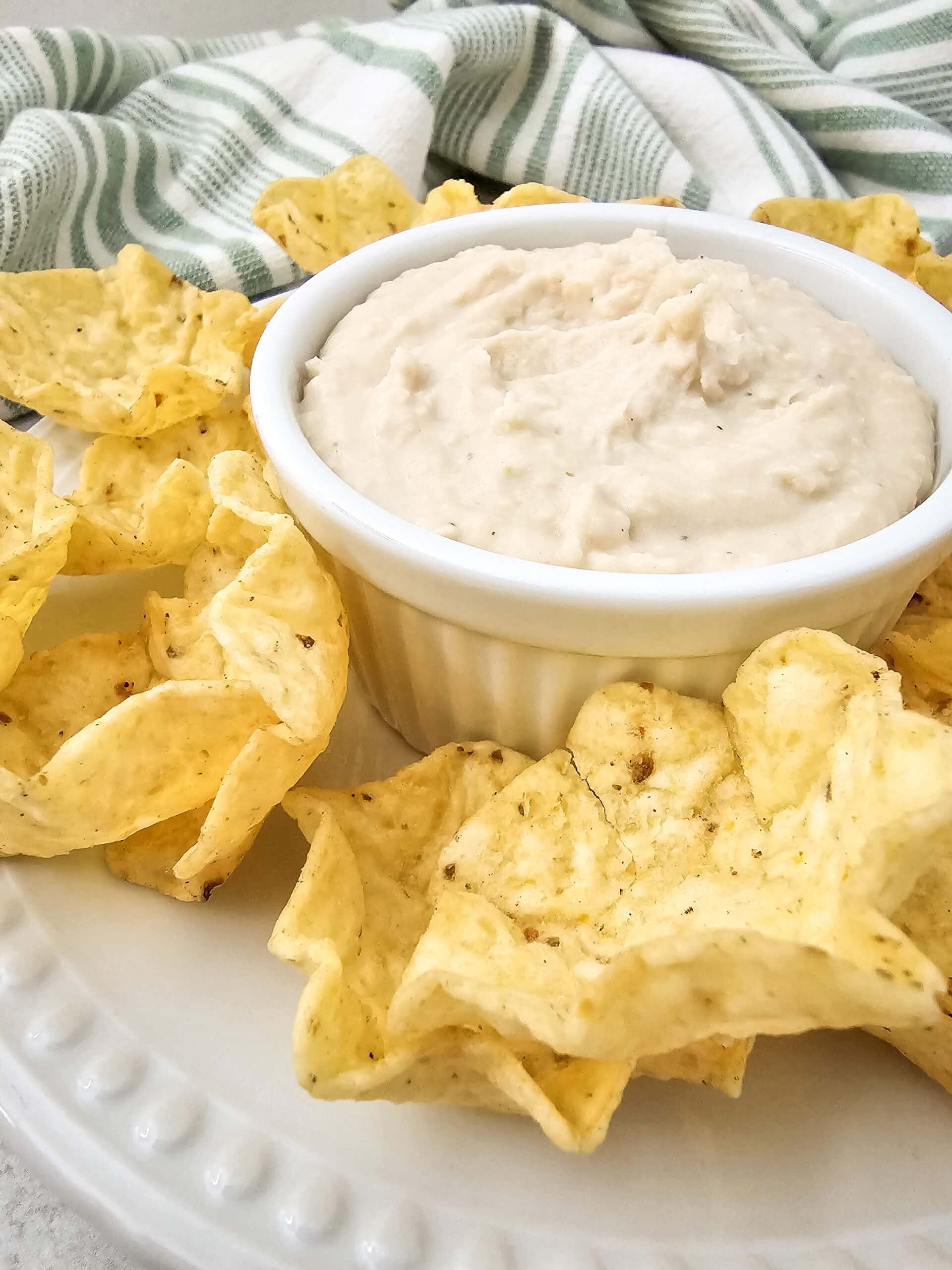 Side view of white bean dip served in a white dish with tortilla chips arranged around it, set on a light surface with a green-striped towel nearby.