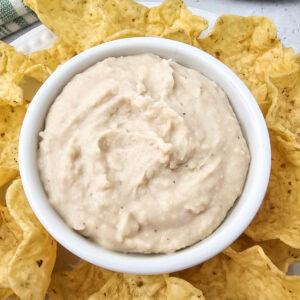Overhead view of white bean dip in a small dish placed on a plate of tortilla chips, ready for dipping.