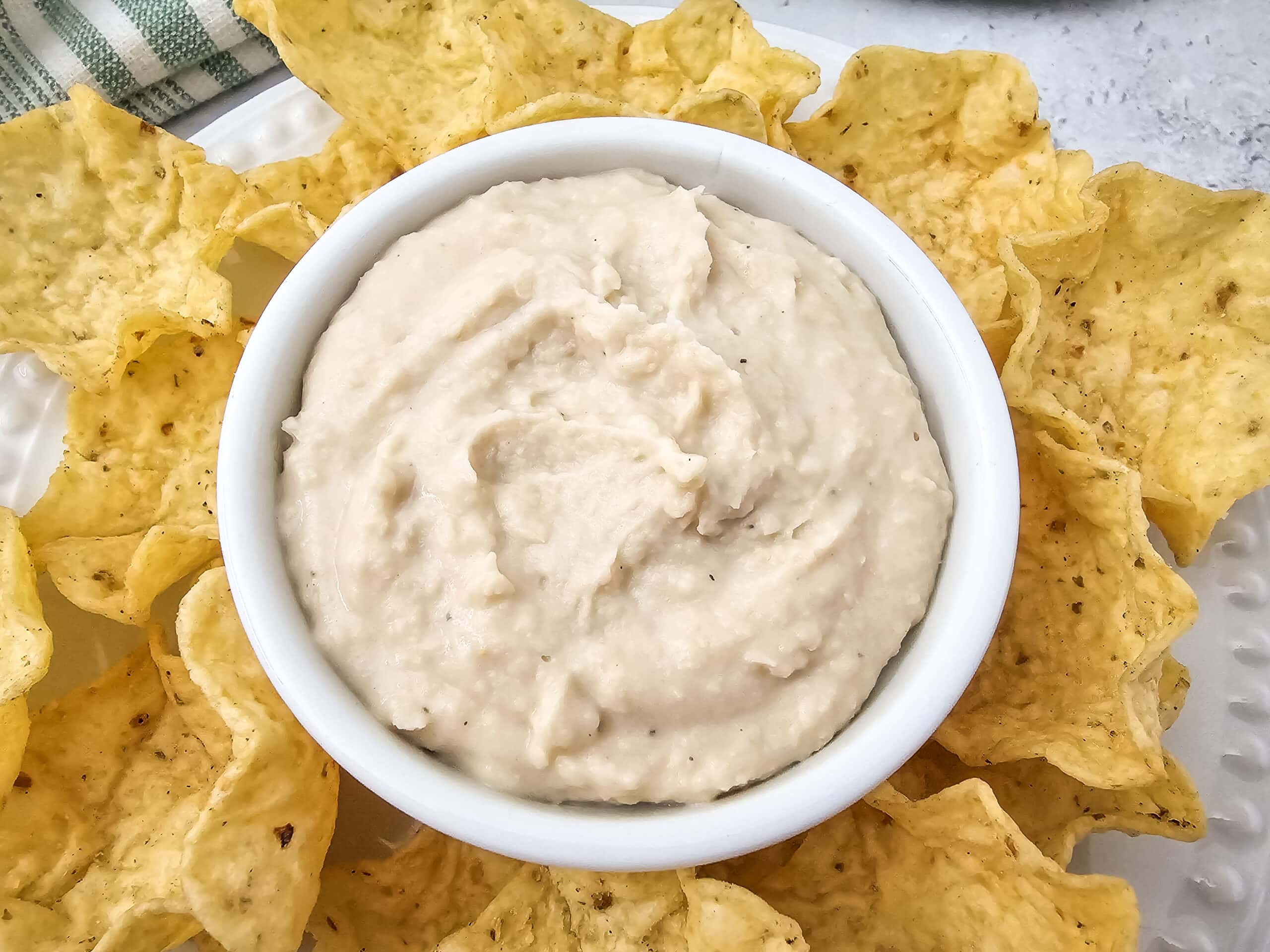 Overhead view of white bean dip in a small dish placed on a plate of tortilla chips, ready for dipping.