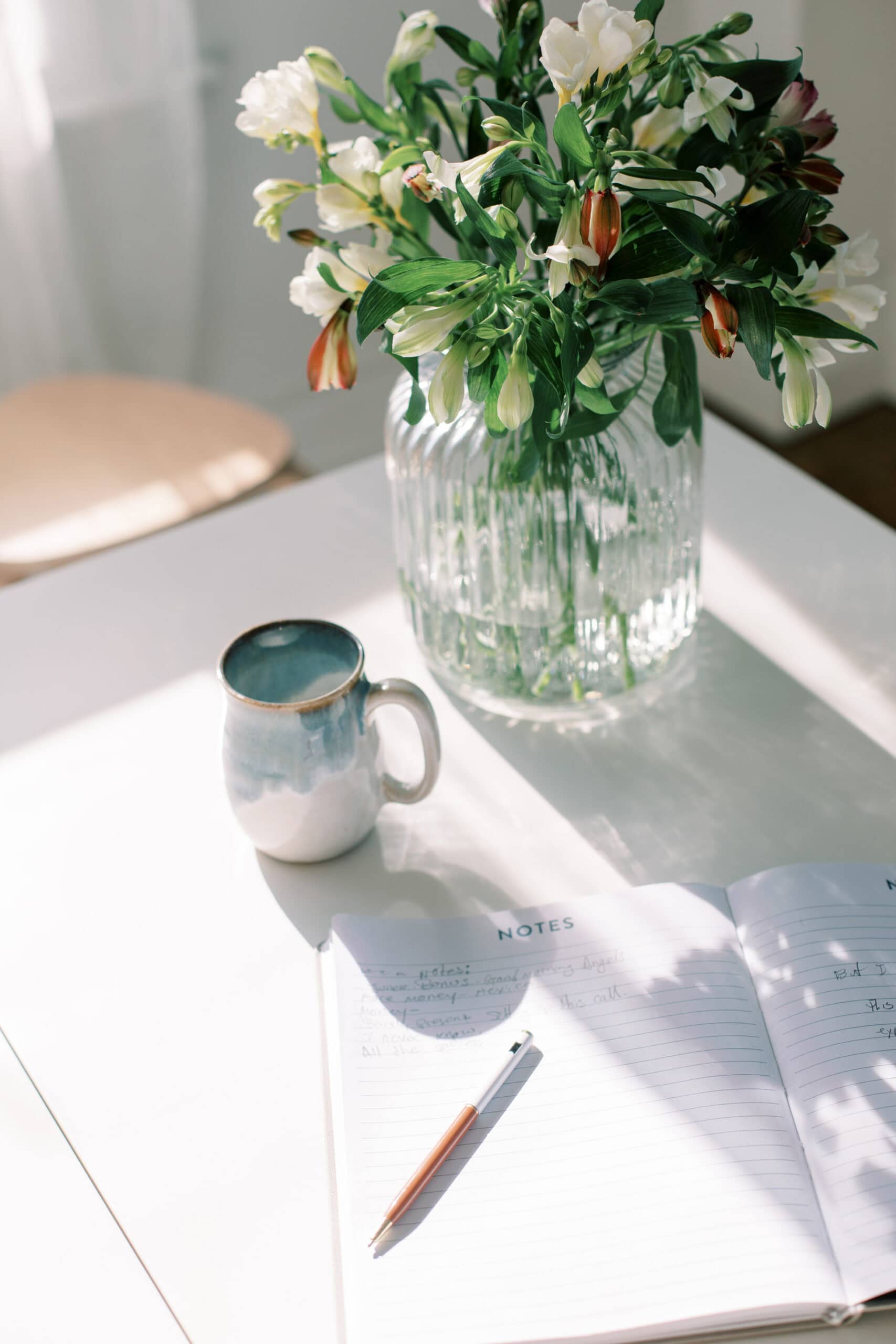 A white table with a clear glass vase of fresh flowers, a handmade ceramic mug, and an open notebook with a pen, all bathed in gentle natural sunlight.