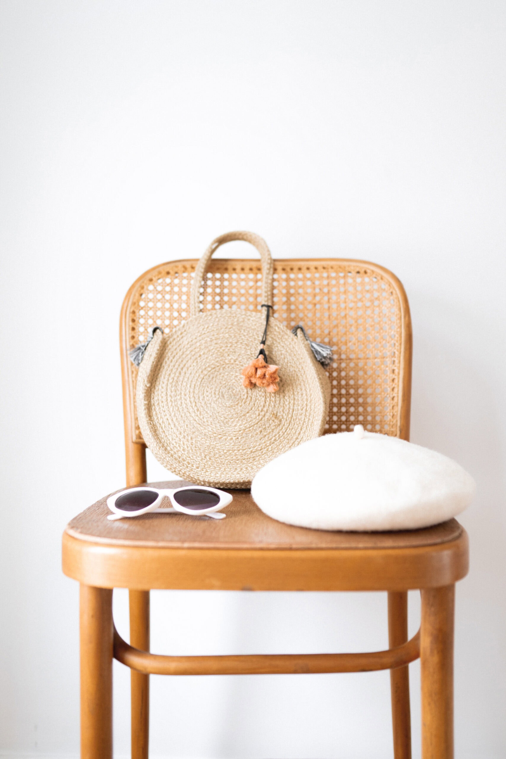A stylish straw bag with a tassel, a pair of white sunglasses, and a cream-colored beret arranged on a woven-back wooden chair against a white background.