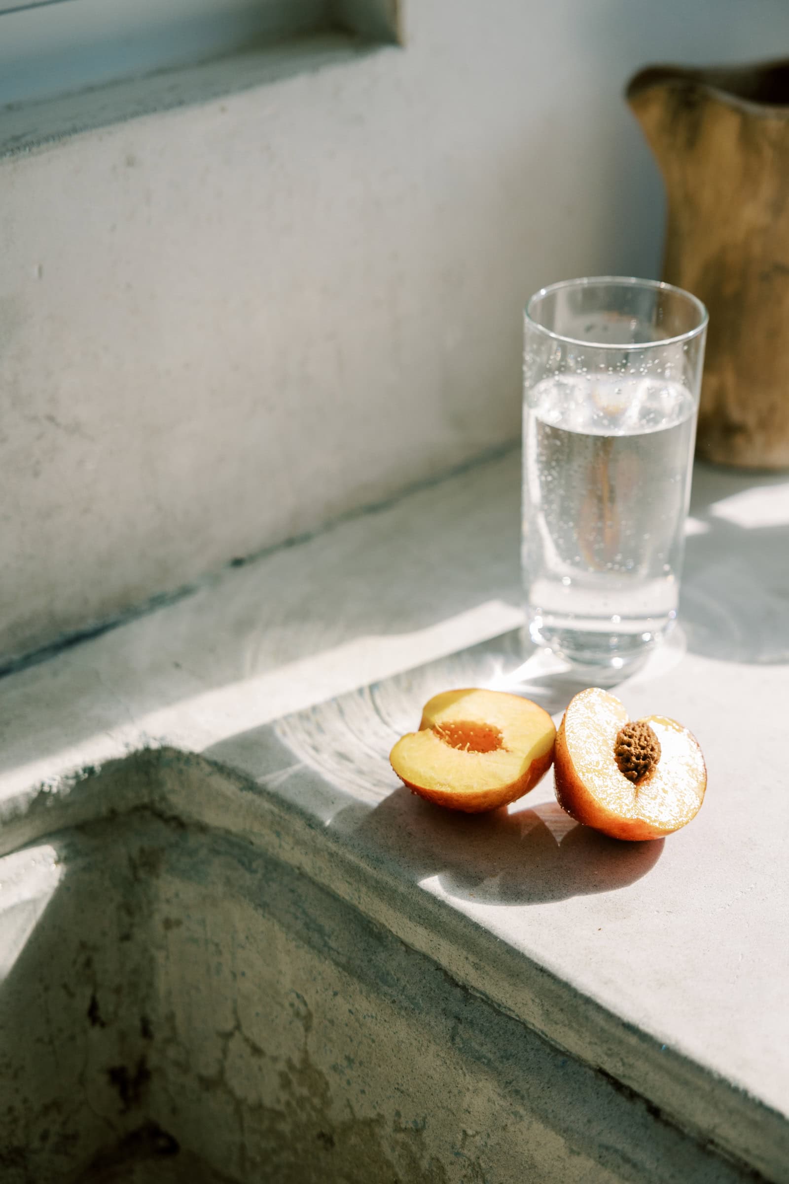 A glass of sparkling water and a fresh peach sliced in half, set on a sunlit stone windowsill, with soft natural light streaming in.