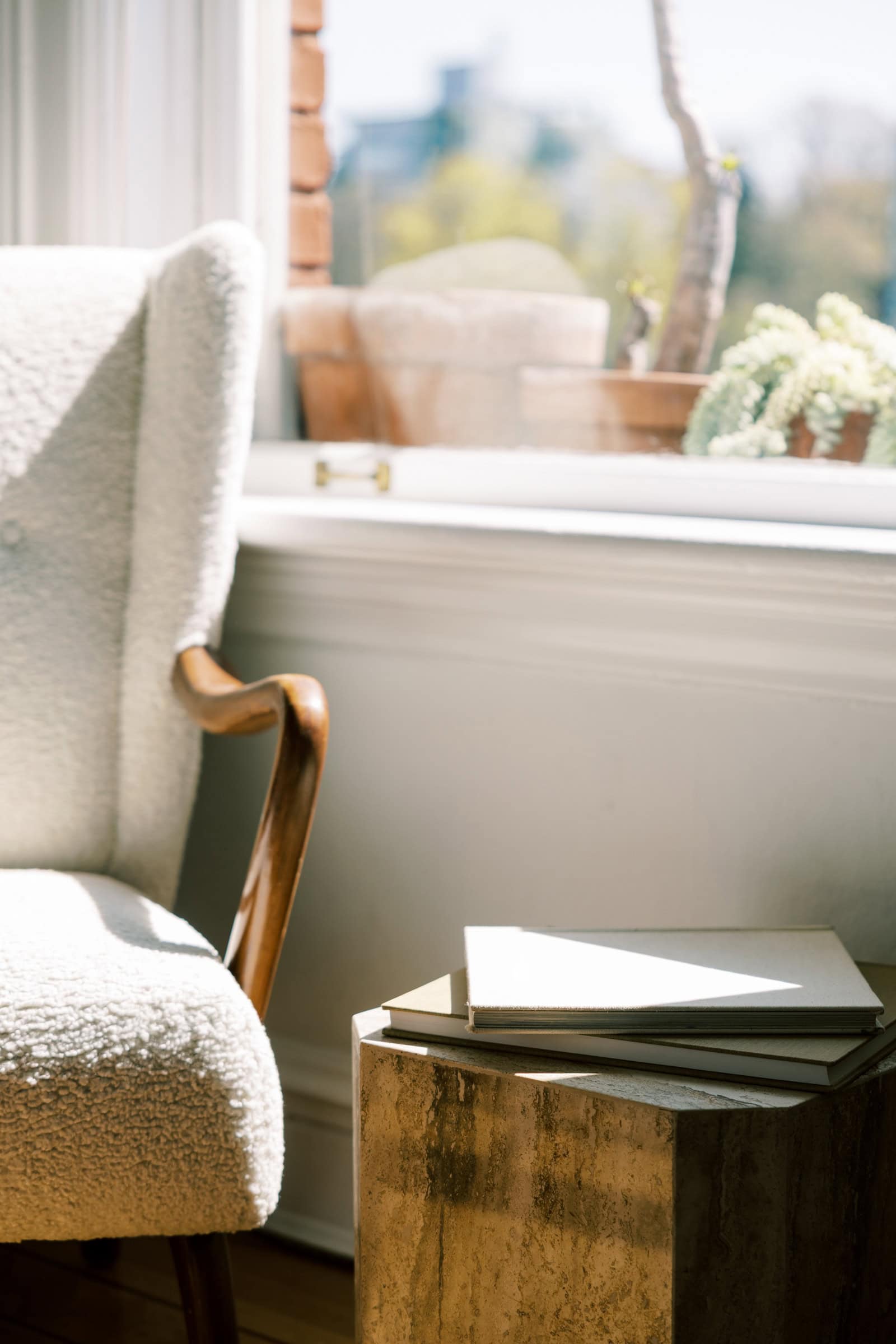 A cozy living space featuring a cream boucle armchair and a small wooden side table with two stacked books, illuminated by sunlight through a window.