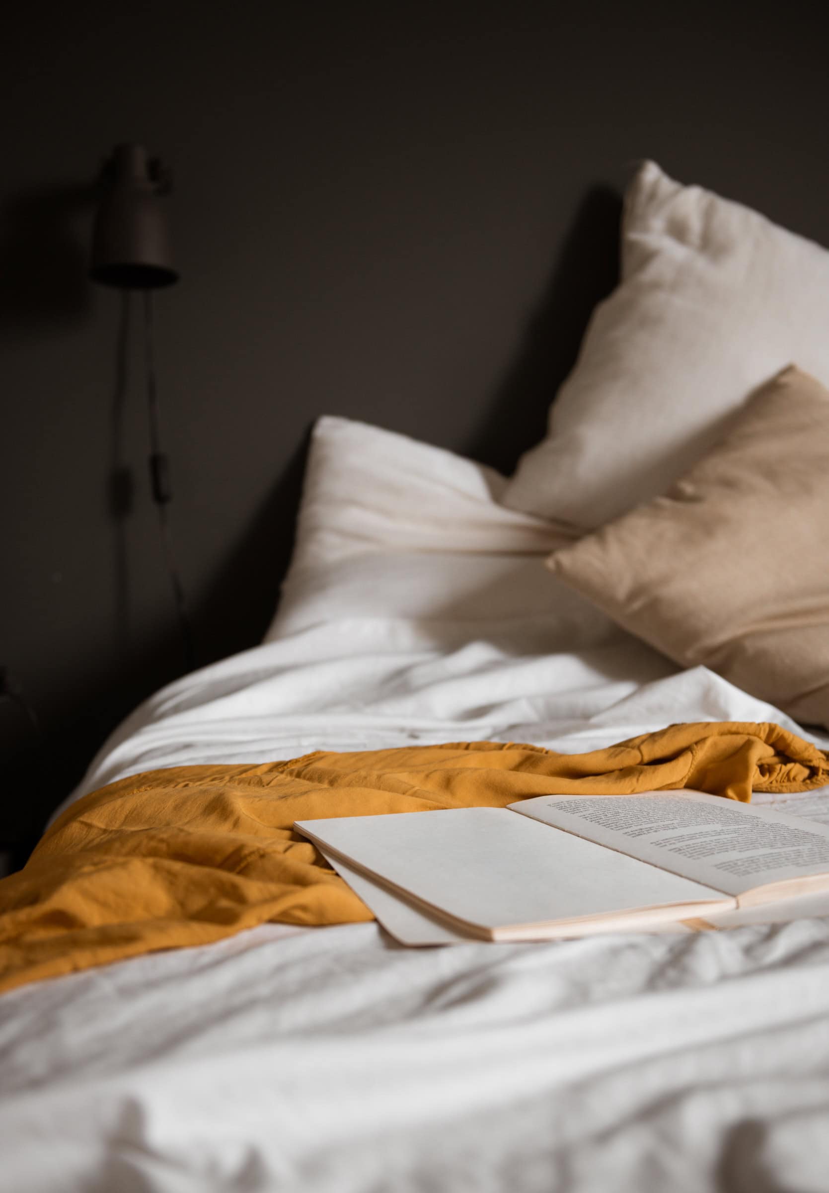 A bed with white and beige pillows and a mustard yellow throw blanket, with an open book resting on top in soft, moody lighting.