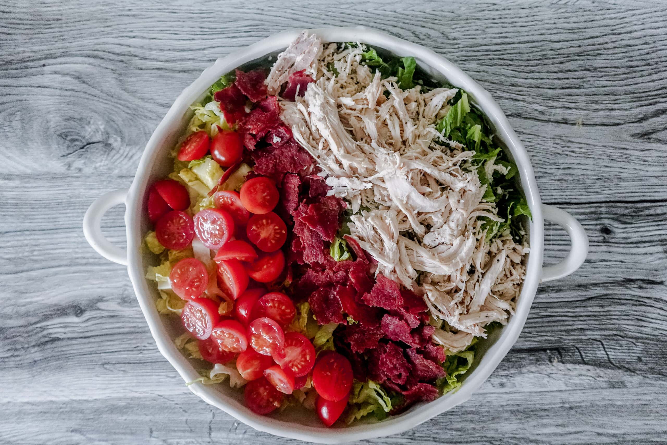Overhead view of a white dish with handles, showing neatly arranged rows of shredded chicken, crispy bacon, and halved cherry tomatoes over chopped lettuce.