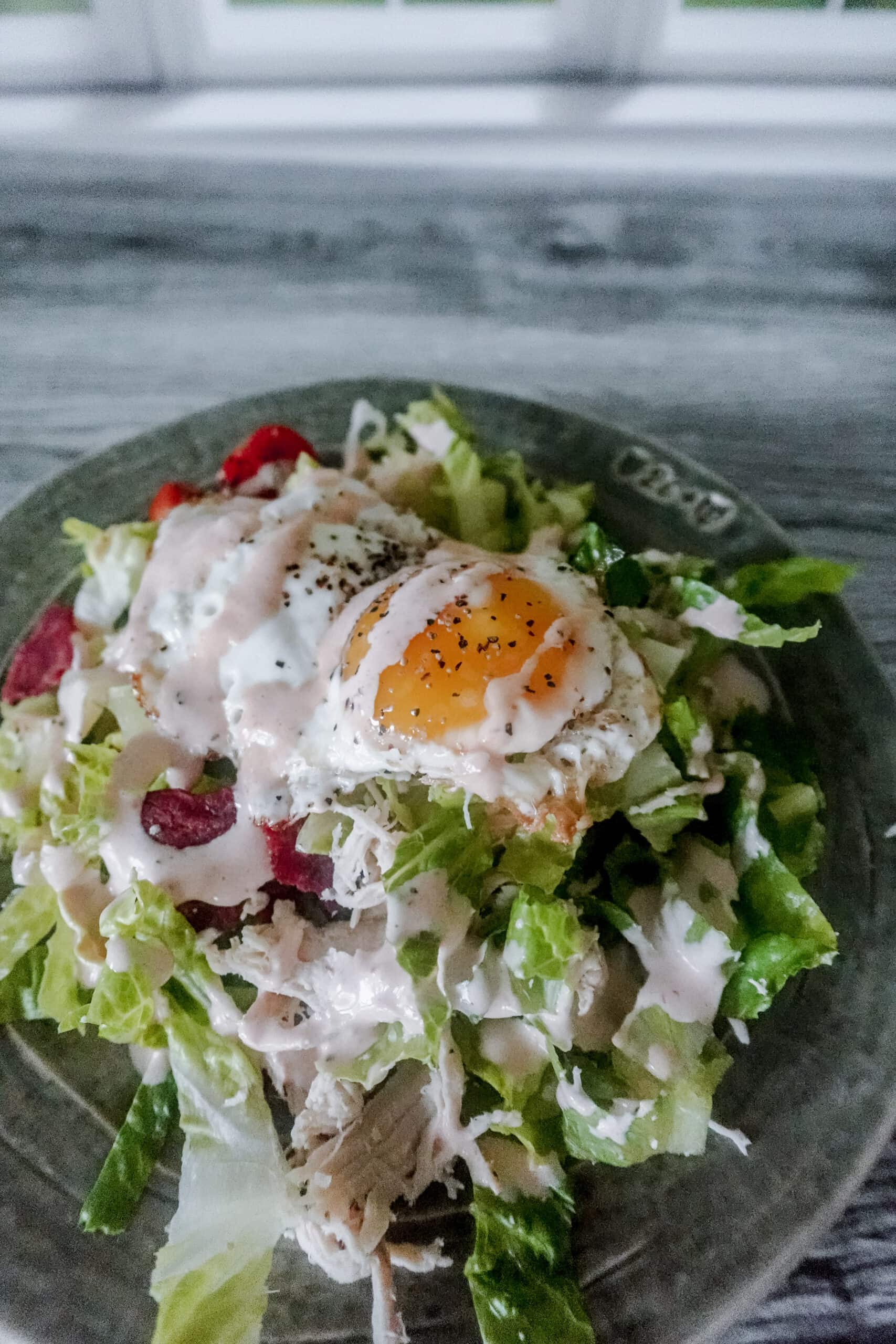 A fried egg with runny yolk sits on top of shredded chicken, bacon, lettuce, and dressing for a hearty salad portion.