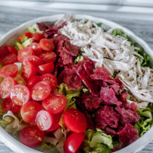 A large white dish brimming with BLT chicken salad, with vibrant cherry tomatoes, crumbled bacon, and shredded chicken arranged over crisp lettuce.