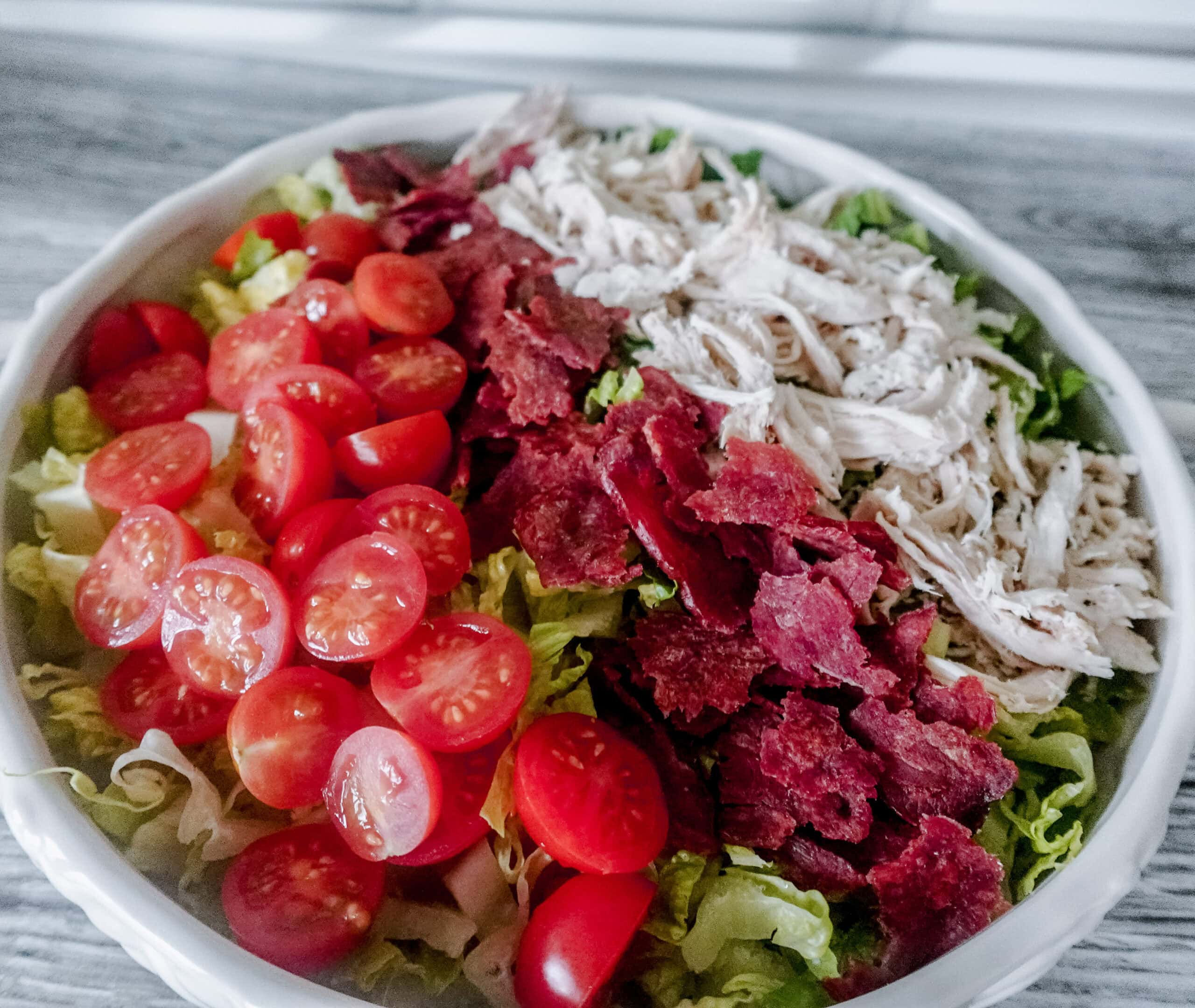 A large white dish brimming with BLT chicken salad, with vibrant cherry tomatoes, crumbled bacon, and shredded chicken arranged over crisp lettuce.