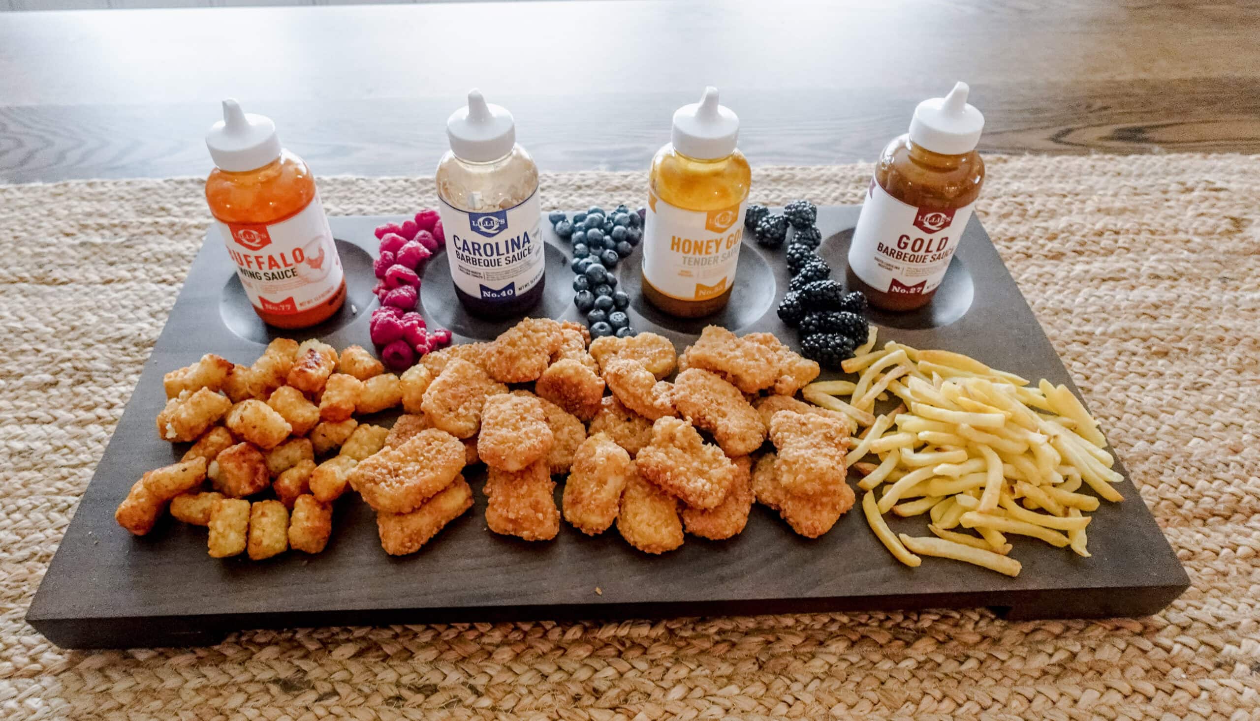 A pile of golden nuggets arranged in front of colorful bottles of buffalo, Carolina barbecue, honey gold, and gold barbecue sauces, with berries as garnish.