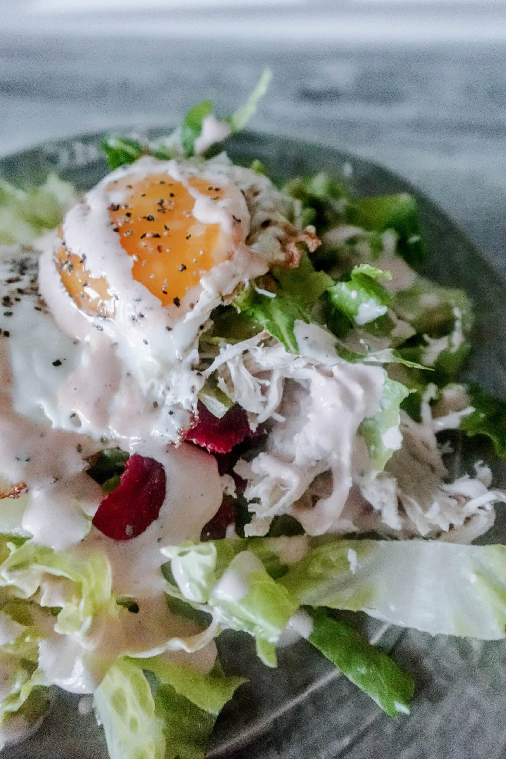 A green plate filled with chopped romaine lettuce, shredded chicken, crispy bacon, cherry tomatoes, and topped with a fried egg, drizzled with creamy BBQ ranch dressing.
