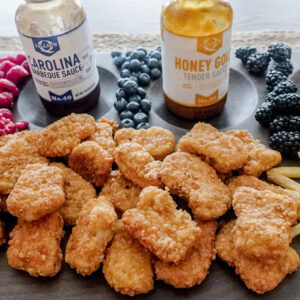 A rectangular serving board with nuggets, fries, tater tots, raspberries, blueberries, blackberries, and four bottles of dipping sauces.