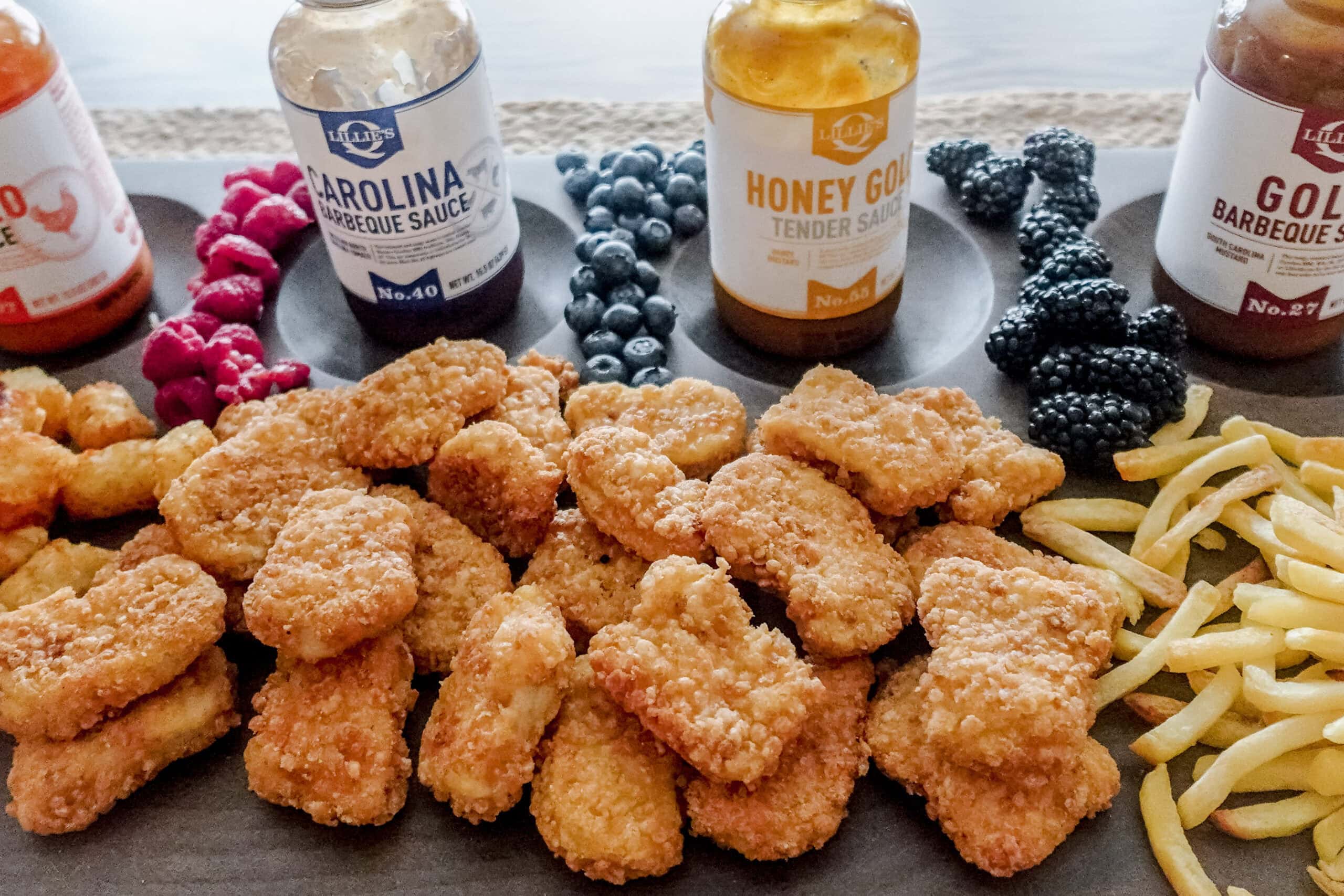 A rectangular serving board with nuggets, fries, tater tots, raspberries, blueberries, blackberries, and four bottles of dipping sauces.