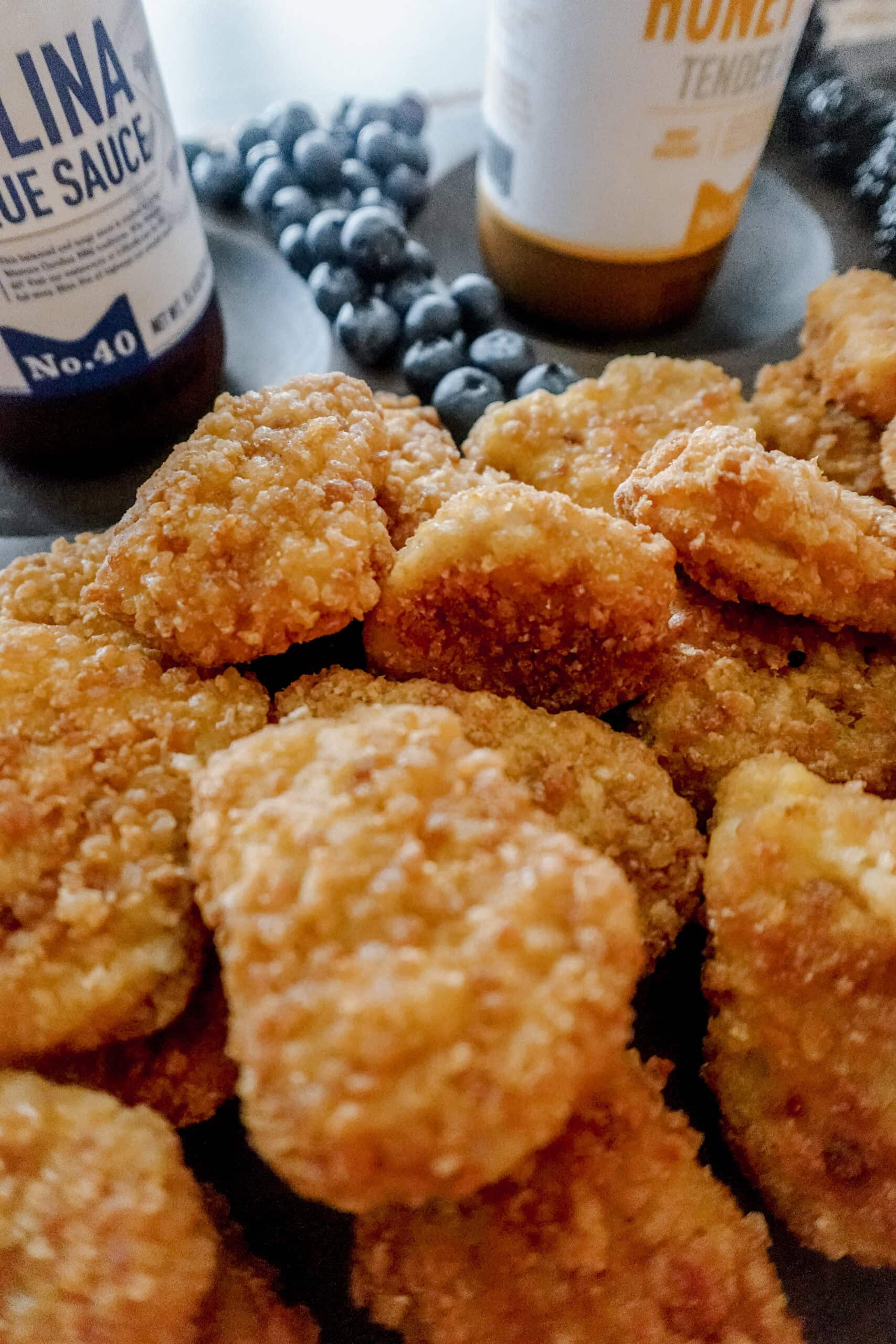Crispy golden brown chicken nuggets stacked in front of dipping sauces, with blueberries in the background.