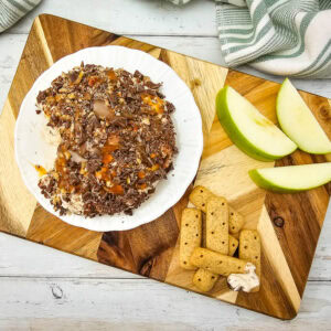 Overhead view of a caramel apple cheeseball on a wooden board with sliced green apples and graham cracker sticks for dipping.