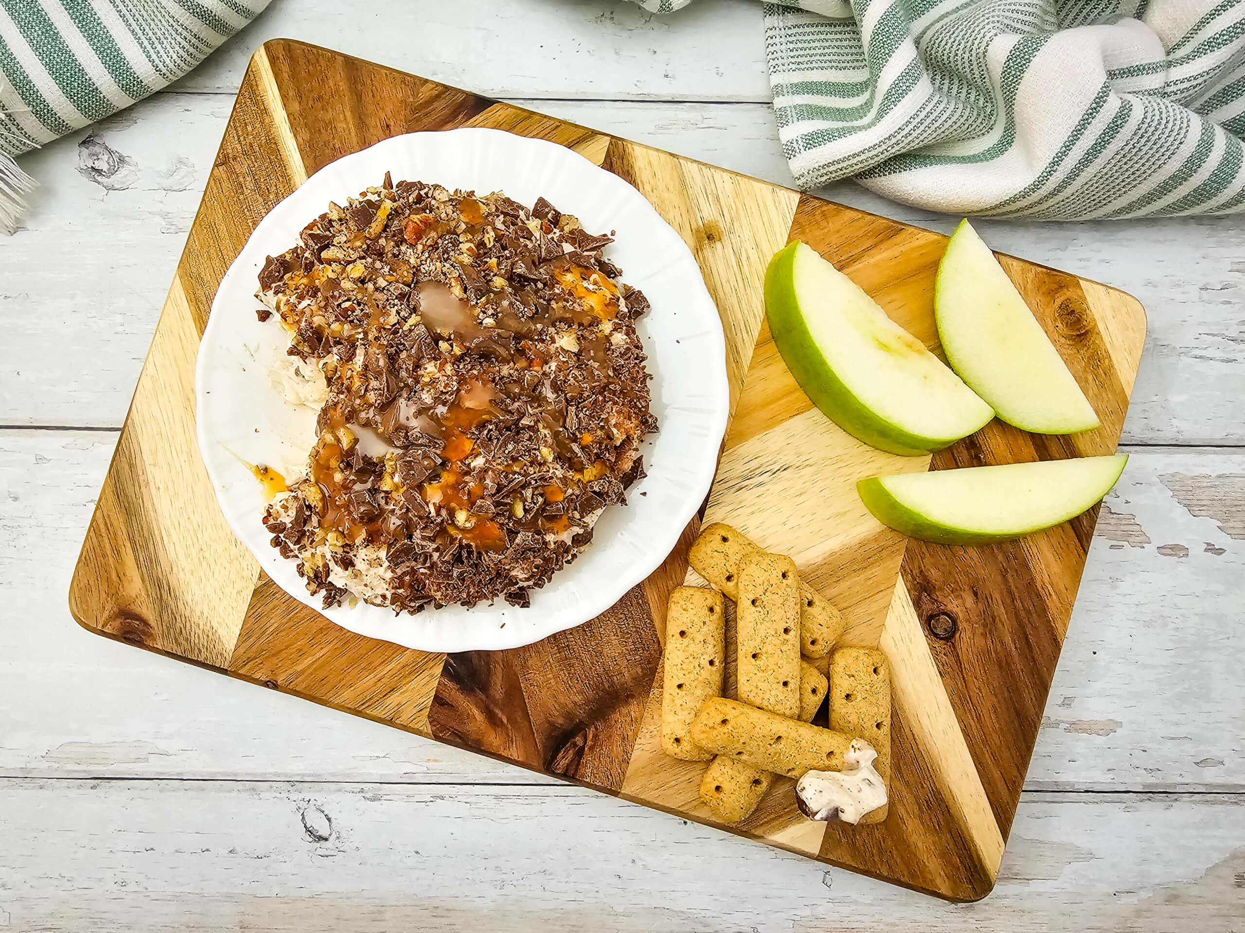 Overhead view of a caramel apple cheeseball on a wooden board with sliced green apples and graham cracker sticks for dipping.