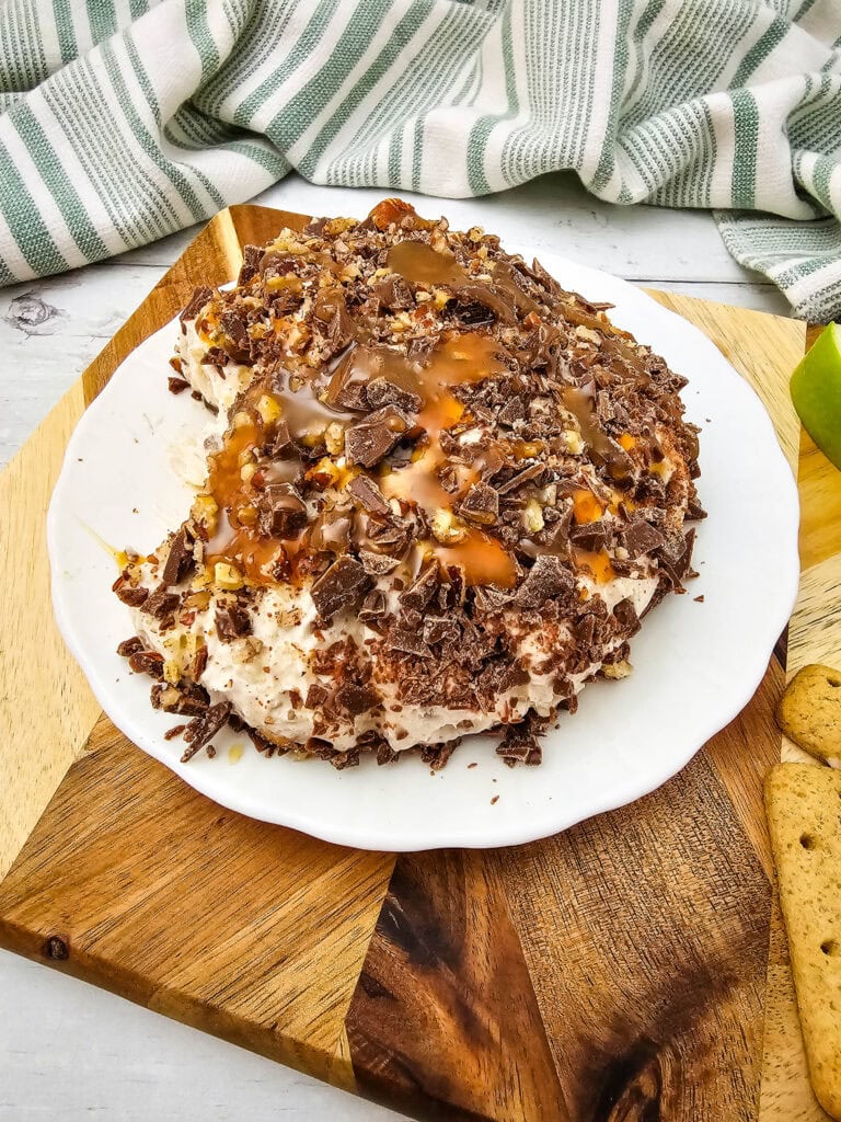 A caramel apple cheeseball covered in chopped chocolate and nuts, topped with caramel drizzle, presented on a white plate.