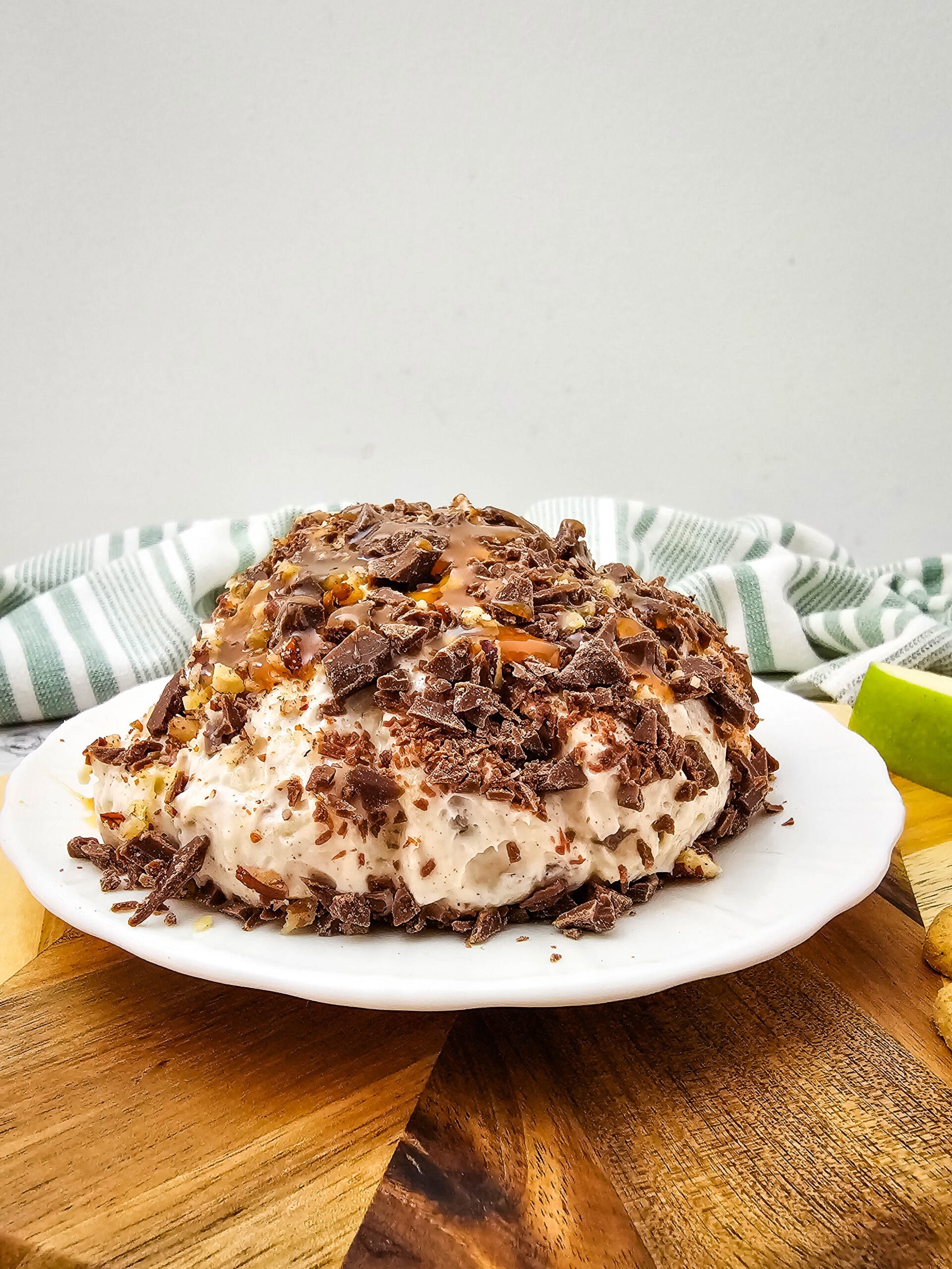A dessert cheeseball coated with chocolate shavings and caramel drizzle, set on a wooden board with apple slices and crackers.
