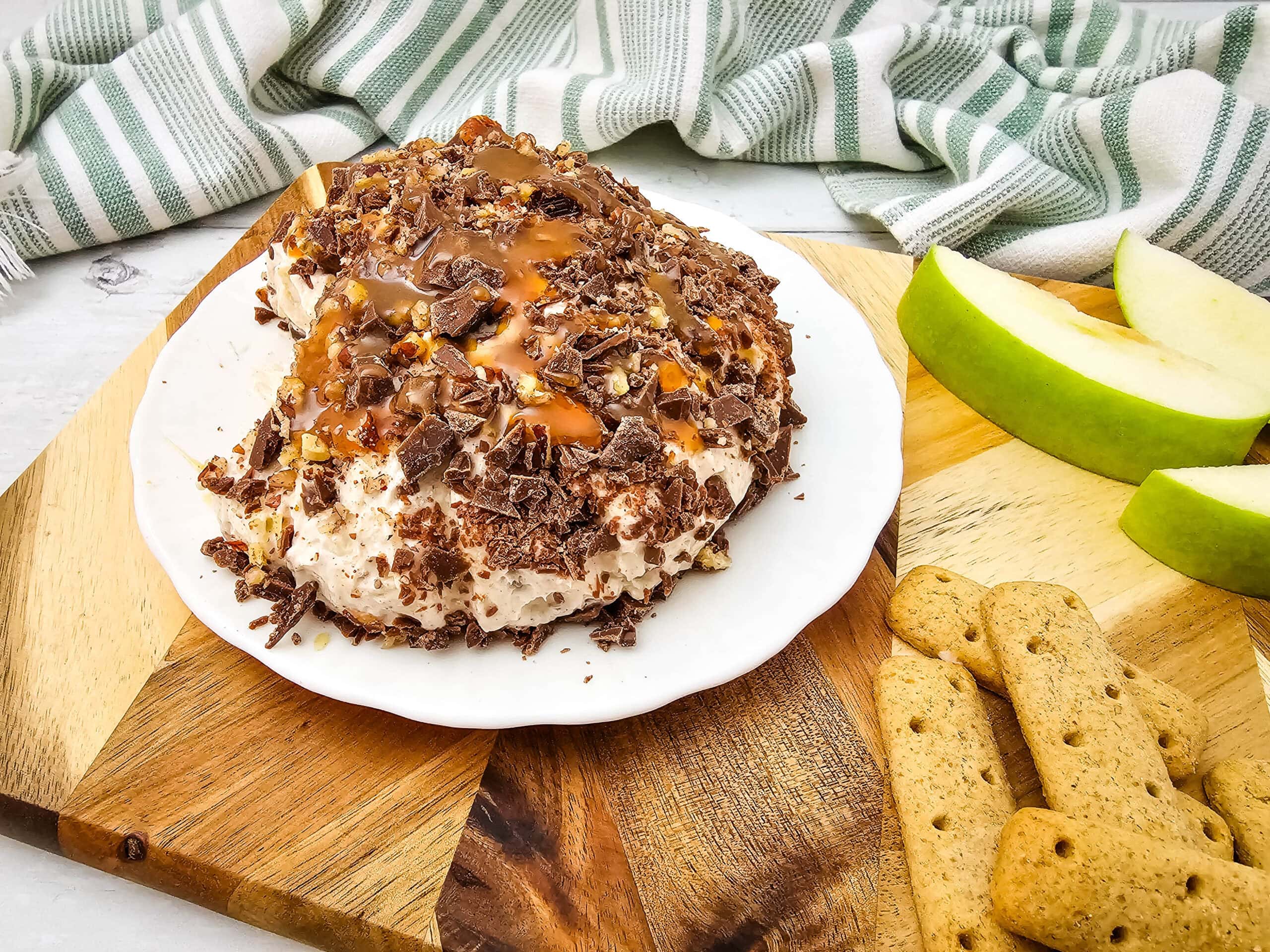 The cheeseball sits on a white plate with chocolate, nuts, and caramel, next to apple slices and graham cracker sticks on a wooden board.