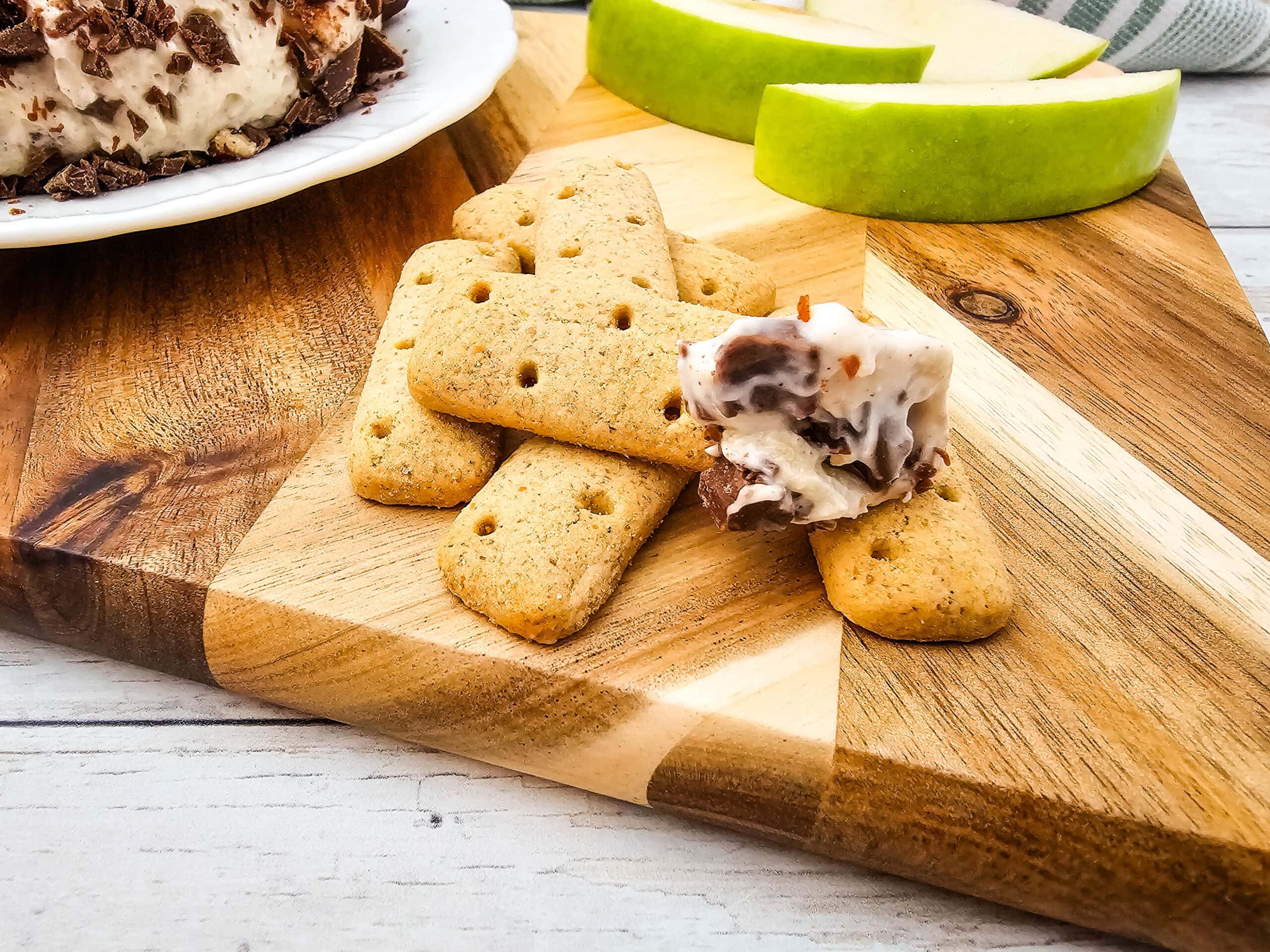 A graham cracker stick topped with creamy caramel apple cheeseball mixture, placed next to more crackers and apple slices on a wooden board.