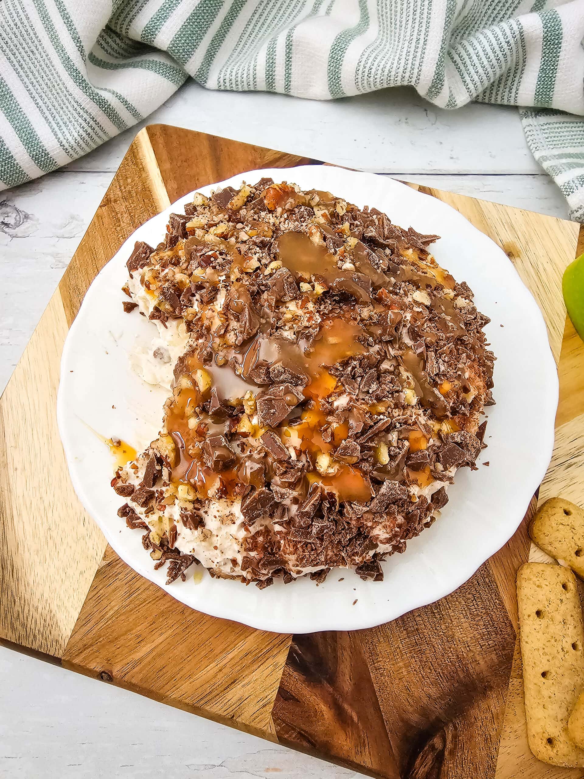 A round cheeseball coated in crushed chocolate, nuts, and drizzled with caramel sauce, served on a white plate.