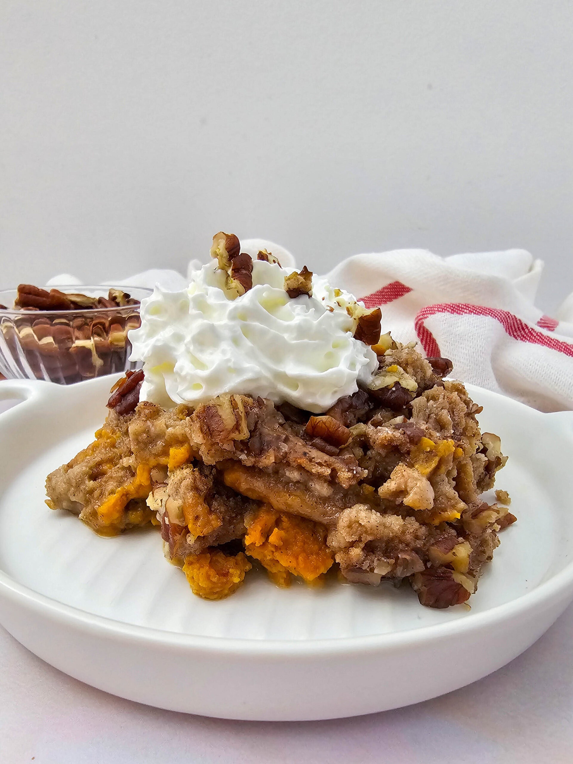 A serving of crockpot pumpkin dump cake topped with whipped cream and chopped pecans on a white plate.