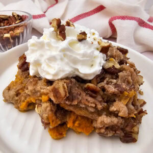 A slice of crockpot pumpkin dump cake topped with whipped cream and pecans, with a bowl of pecans in the background.