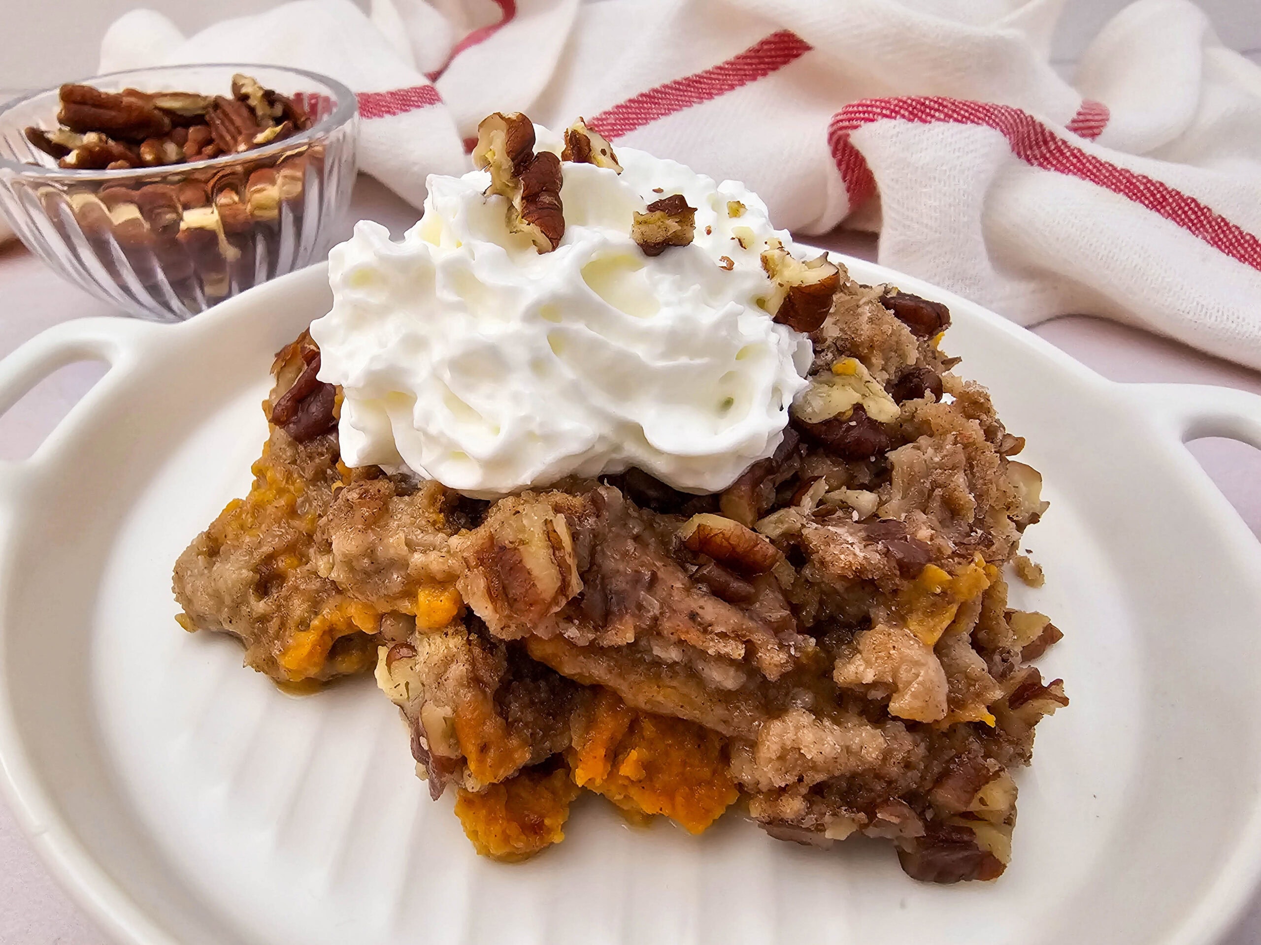 A slice of crockpot pumpkin dump cake topped with whipped cream and pecans, with a bowl of pecans in the background.