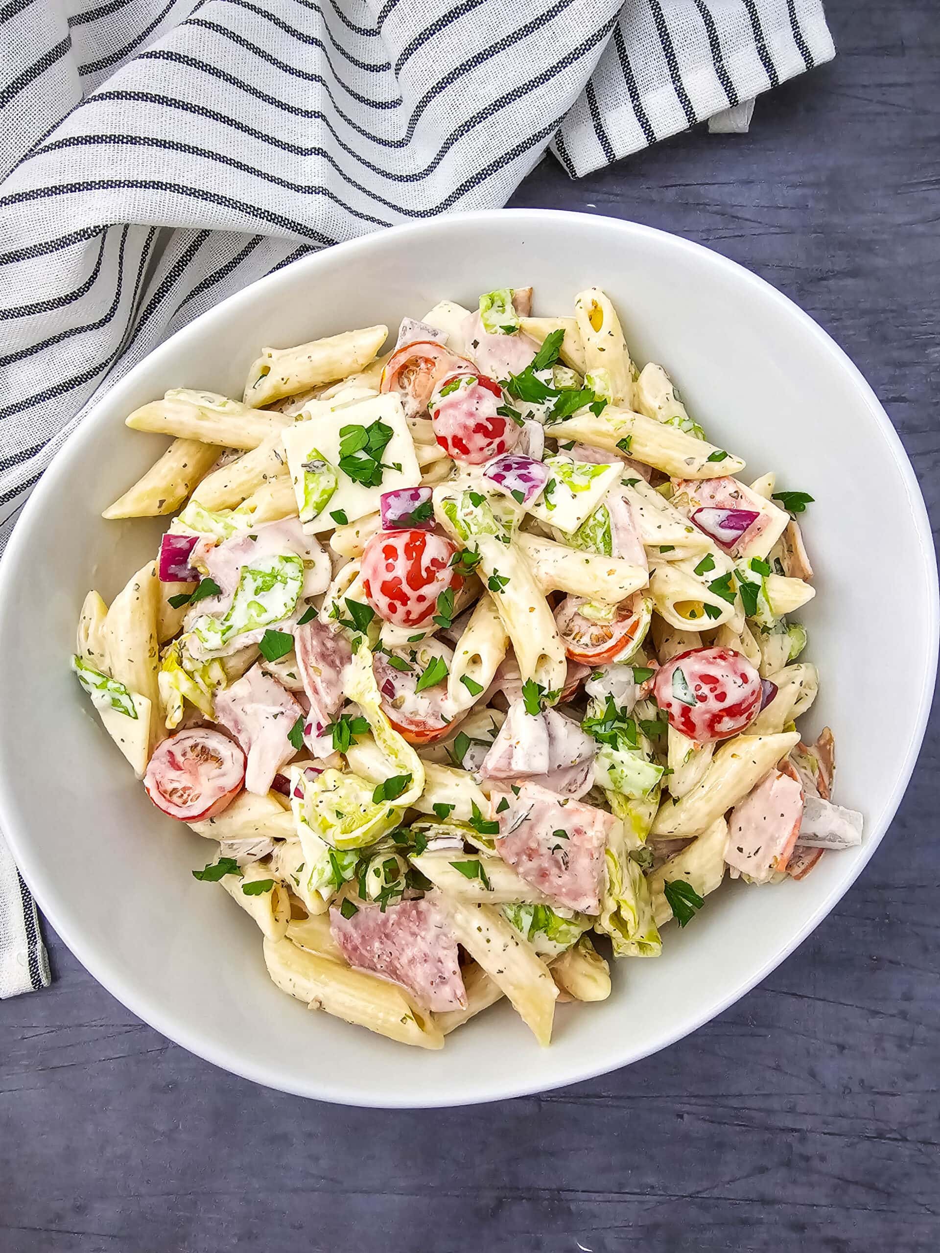 A white bowl filled with pasta salad featuring salami slices, bright cherry tomatoes, red onion, banana peppers, and parsley garnish.