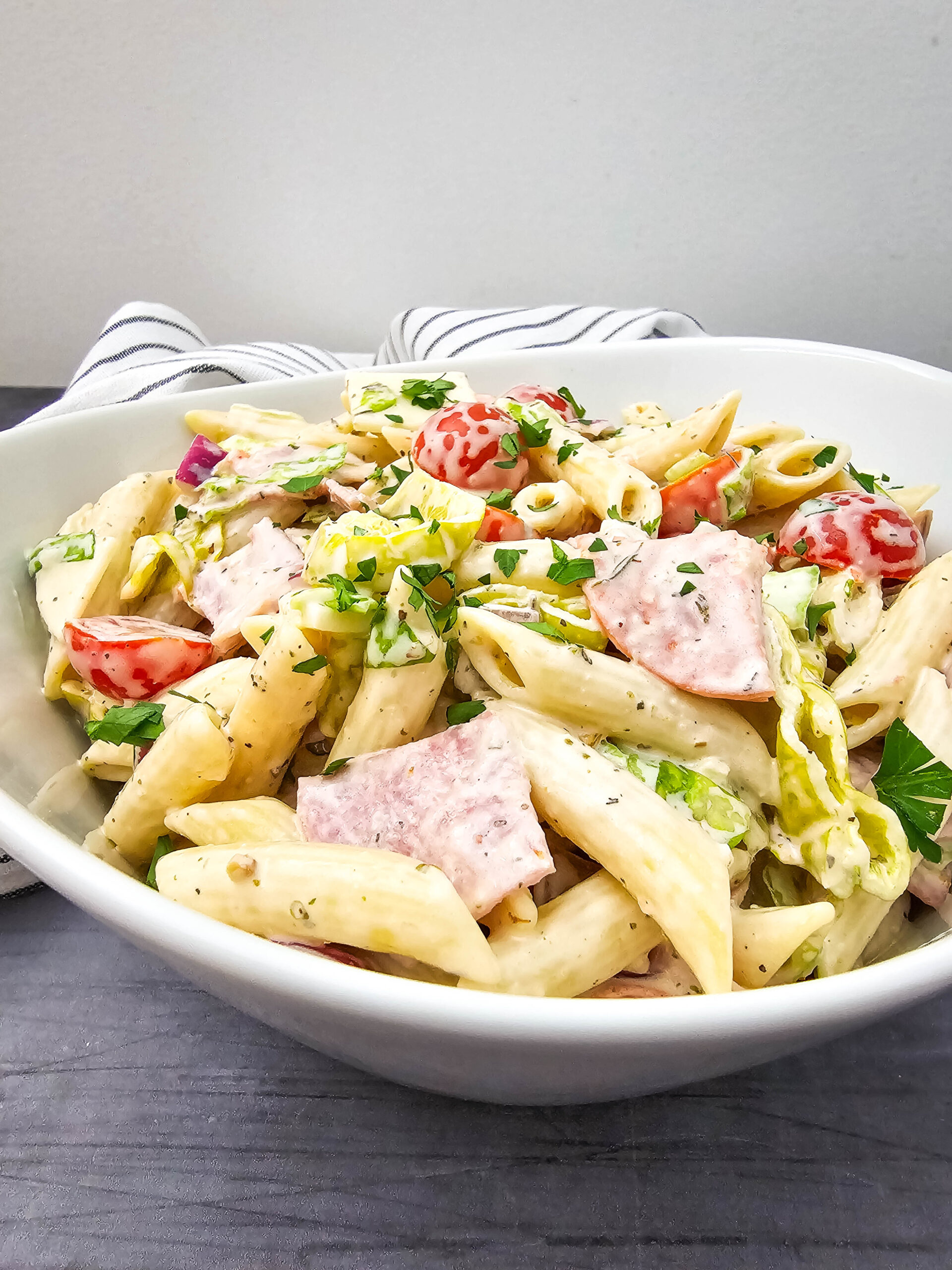 A creamy pasta salad loaded with colorful ingredients like tomatoes, deli meats, cheese, and peppers, set against a gray table with a striped towel.