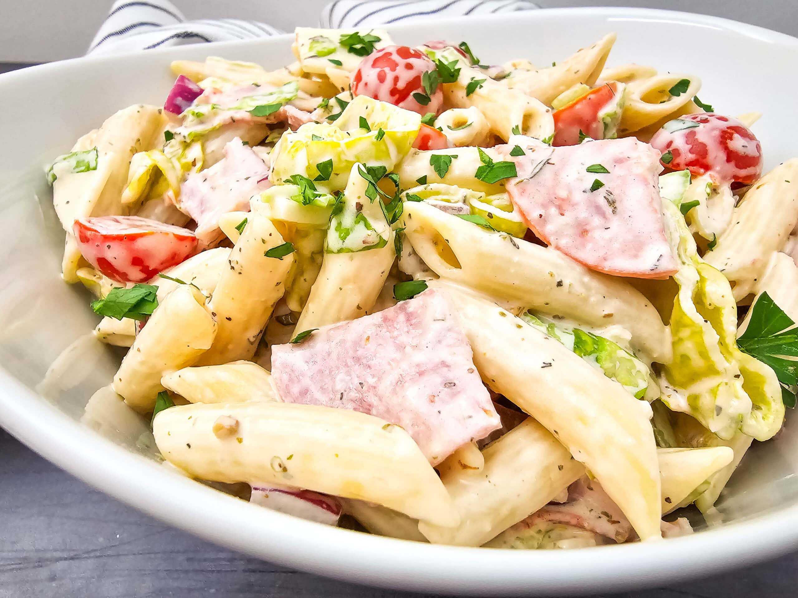 Penne pasta tossed with creamy dressing, cherry tomatoes, salami slices, banana peppers, red onion, and parsley in a white serving bowl.