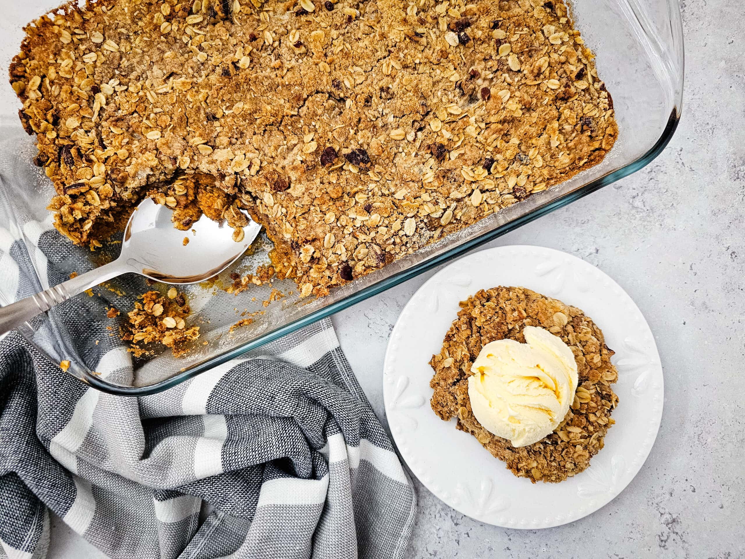 A partially scooped glass dish of pumpkin pie crisp next to a plated serving with vanilla ice cream.