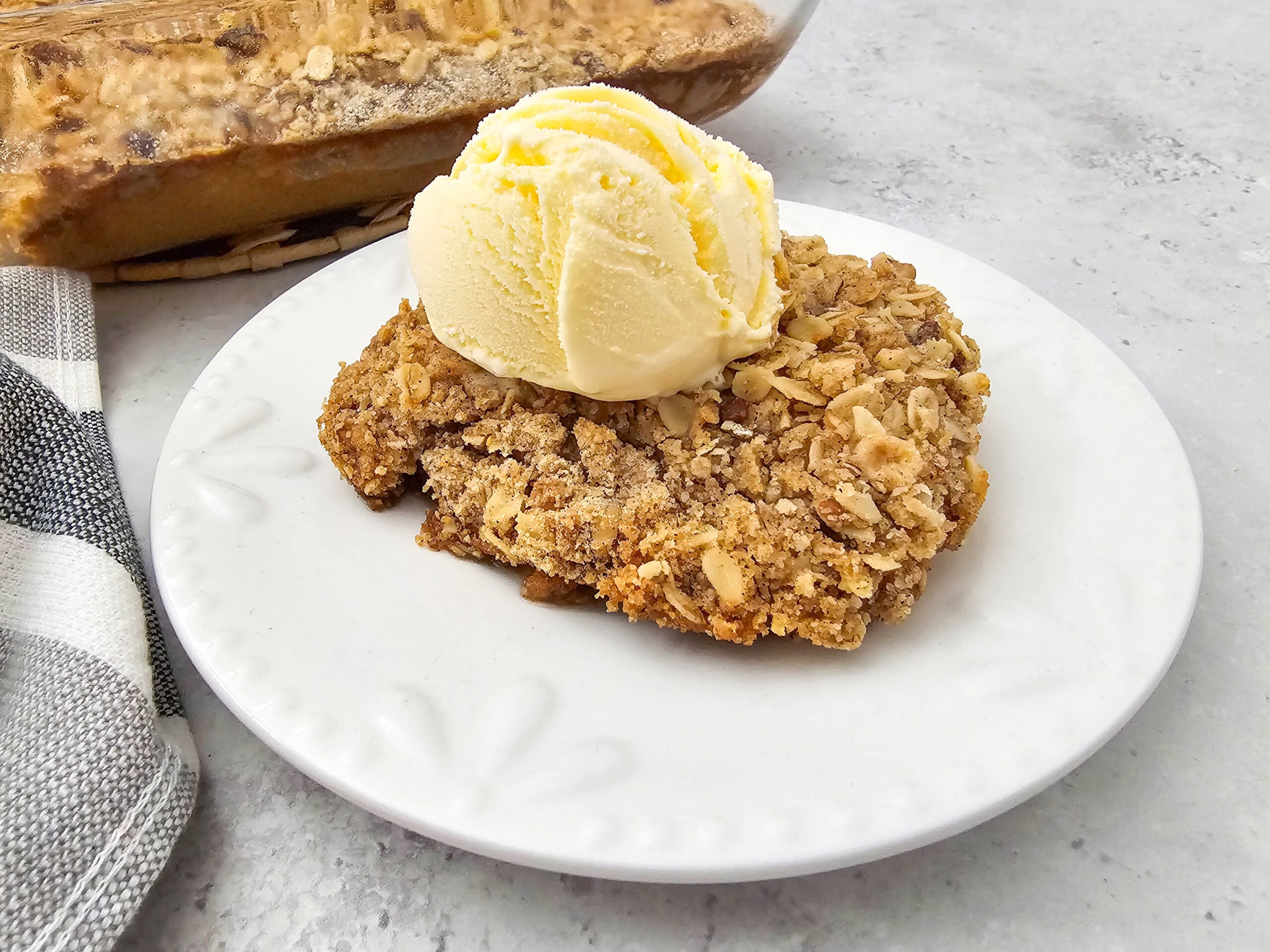 A serving of pumpkin pie crisp topped with a scoop of vanilla ice cream on a white plate, with the baking dish in the background.