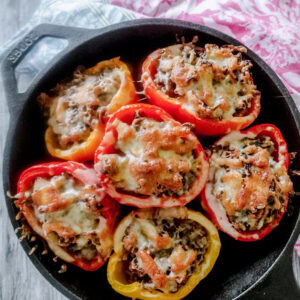 A cast iron skillet is filled with 6 baked stuffed red and yellow bell peppers, topped with melted cheese. The skillet sits on a wooden table with a pink and blue patterned cloth nearby.