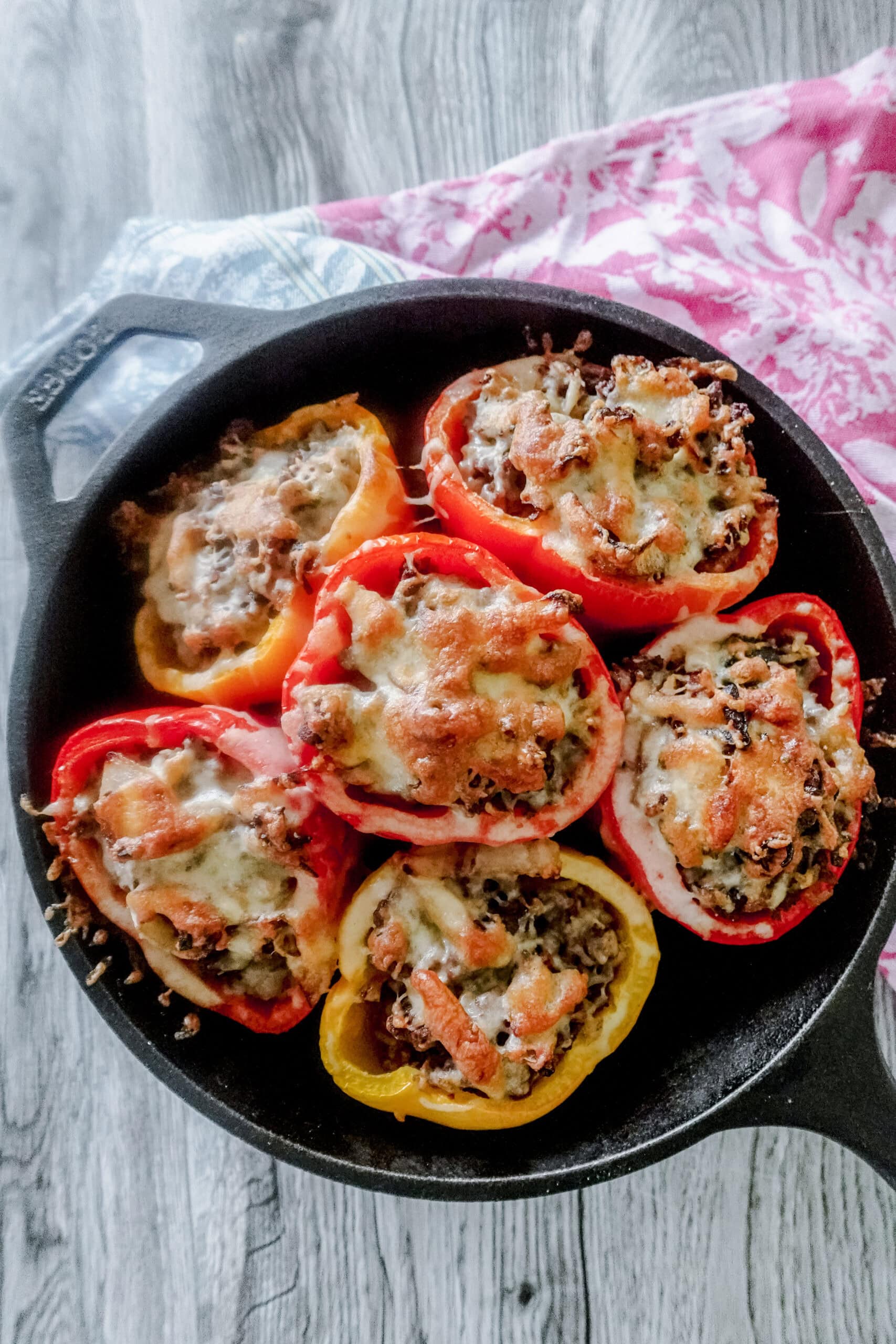 A cast iron skillet is filled with 6 baked stuffed red and yellow bell peppers, topped with melted cheese. The skillet sits on a wooden table with a pink and blue patterned cloth nearby.