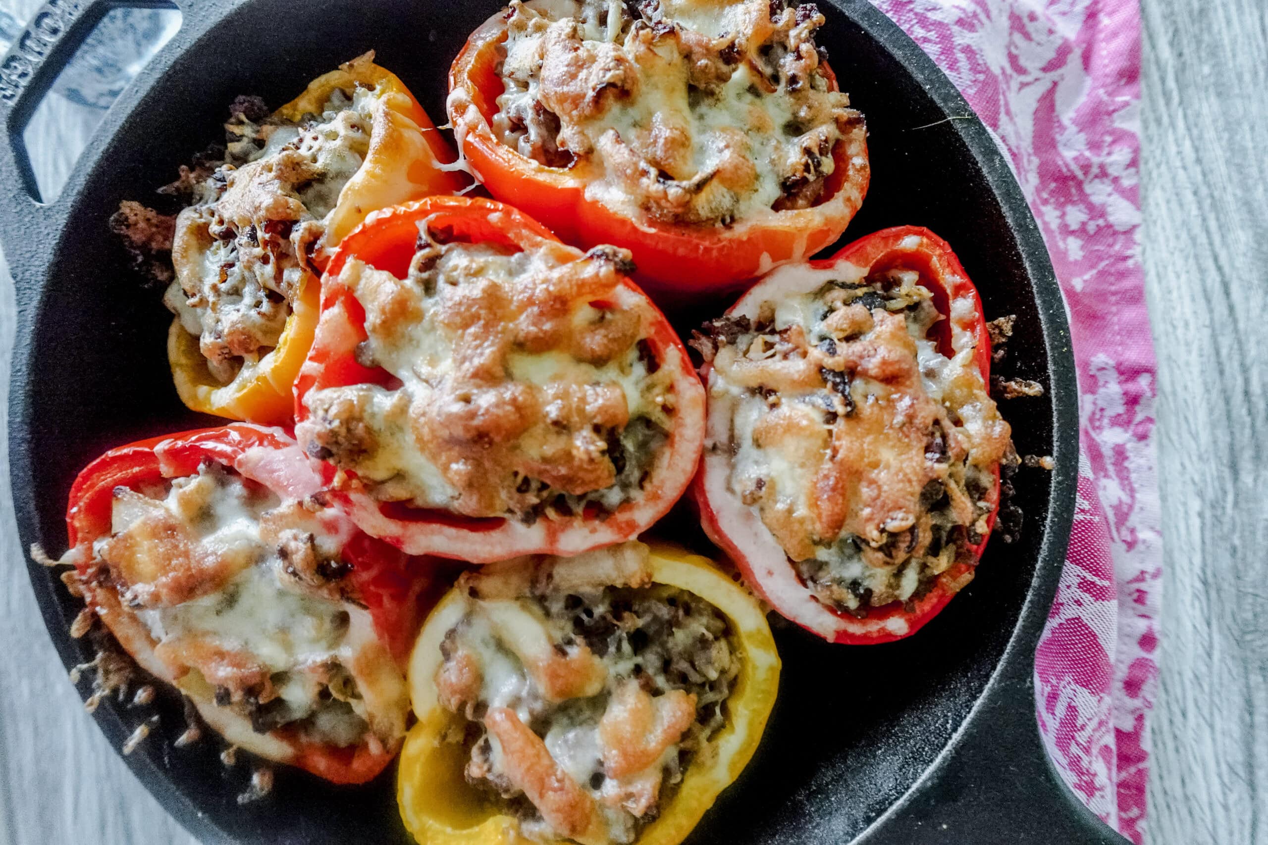 An overhead shot of 6 baked stuffed bell peppers topped with gooey cheese sit in a cast iron skillet on a gray wooden table.