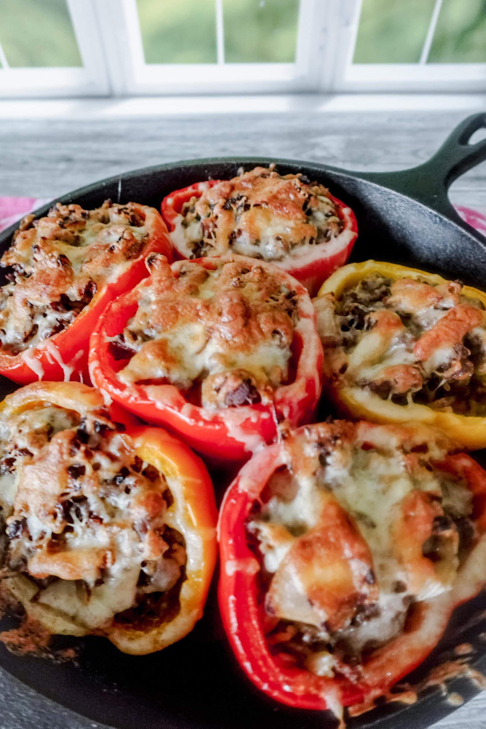 6 baked stuffed bell peppers topped with gooey cheese sit in a cast iron skillet on a gray wooden table. There is a window out of focus in the background.